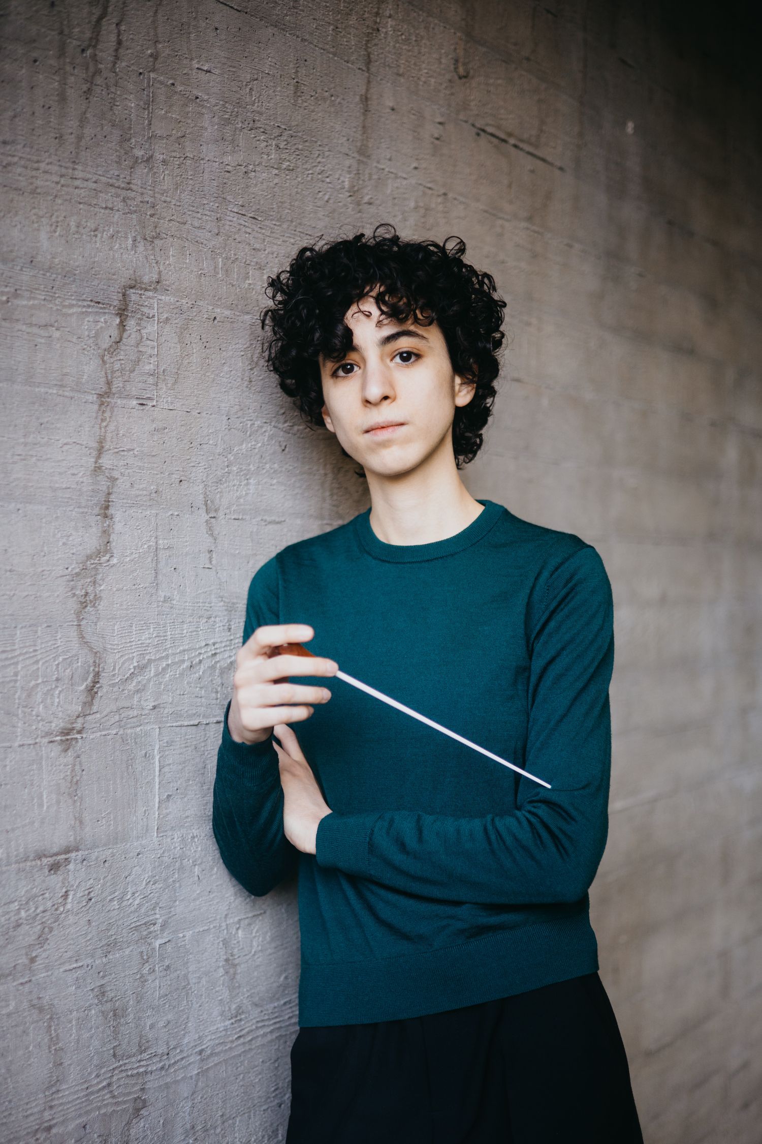 Young male conductor with curly hair holds a baton while leaning against a concrete wall.