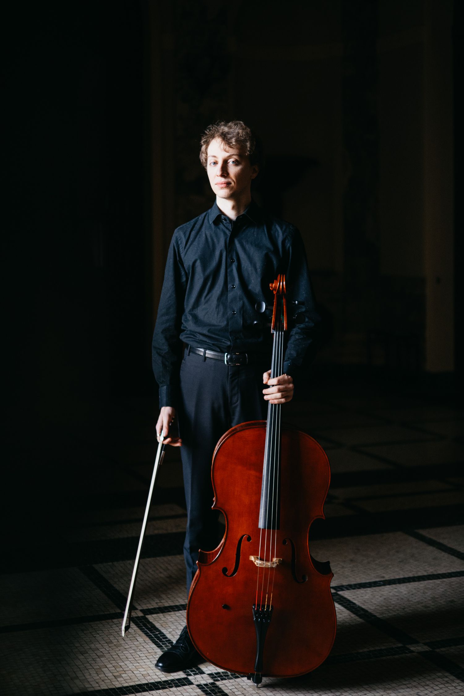Male cellist in black shirt poses with cello and bow in moody lighting.