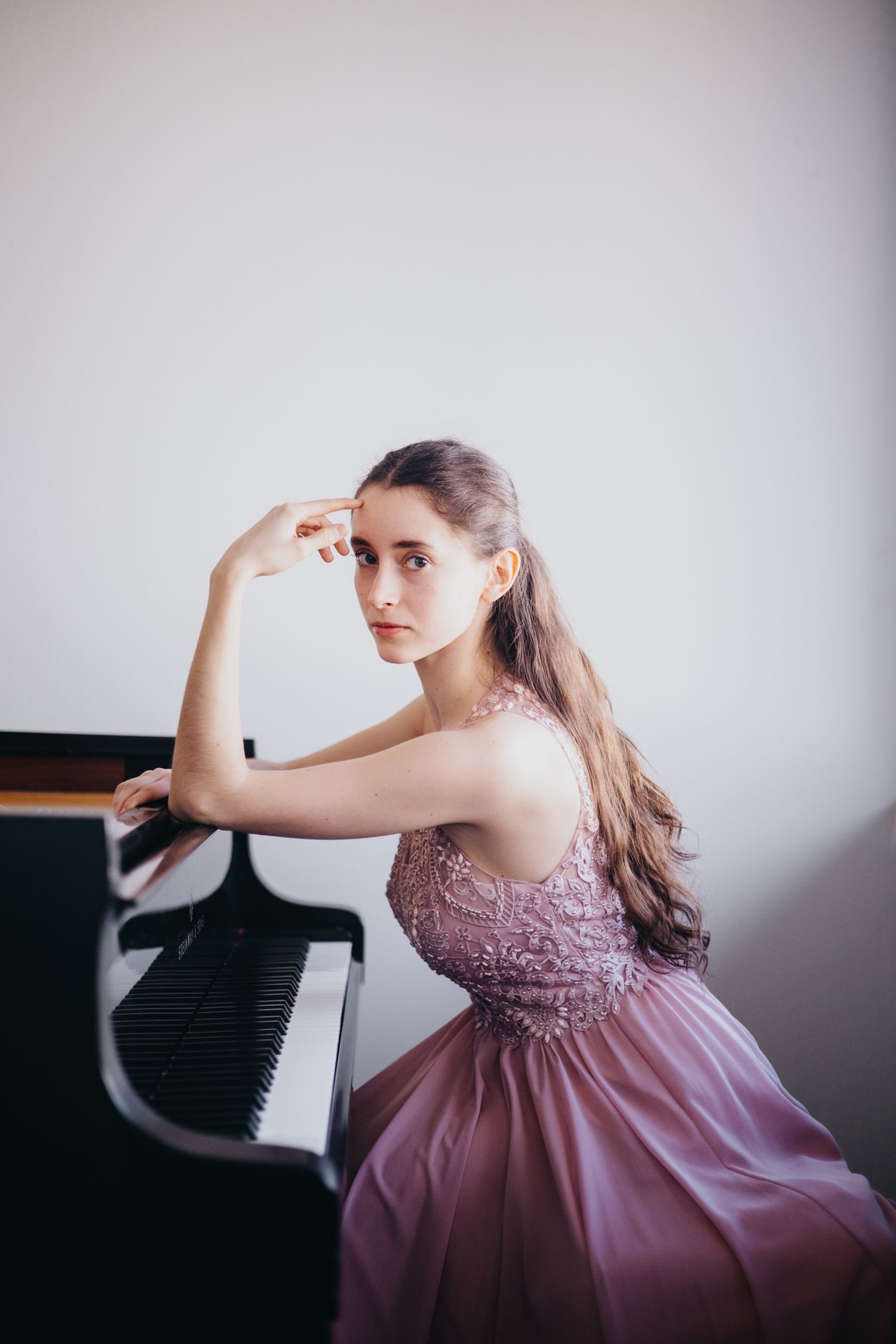 Young woman in pink lace gown posing gracefully beside a grand piano in an elegant studio setting.