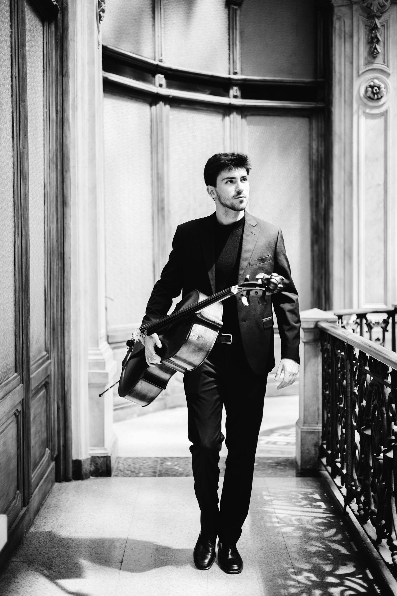 Young male cellist in black suit carrying cello through ornate classical building corridor, black and white photo.