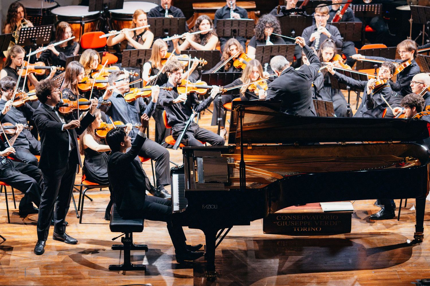 Pianist performing with full orchestra on a concert hall stage under bright lights.