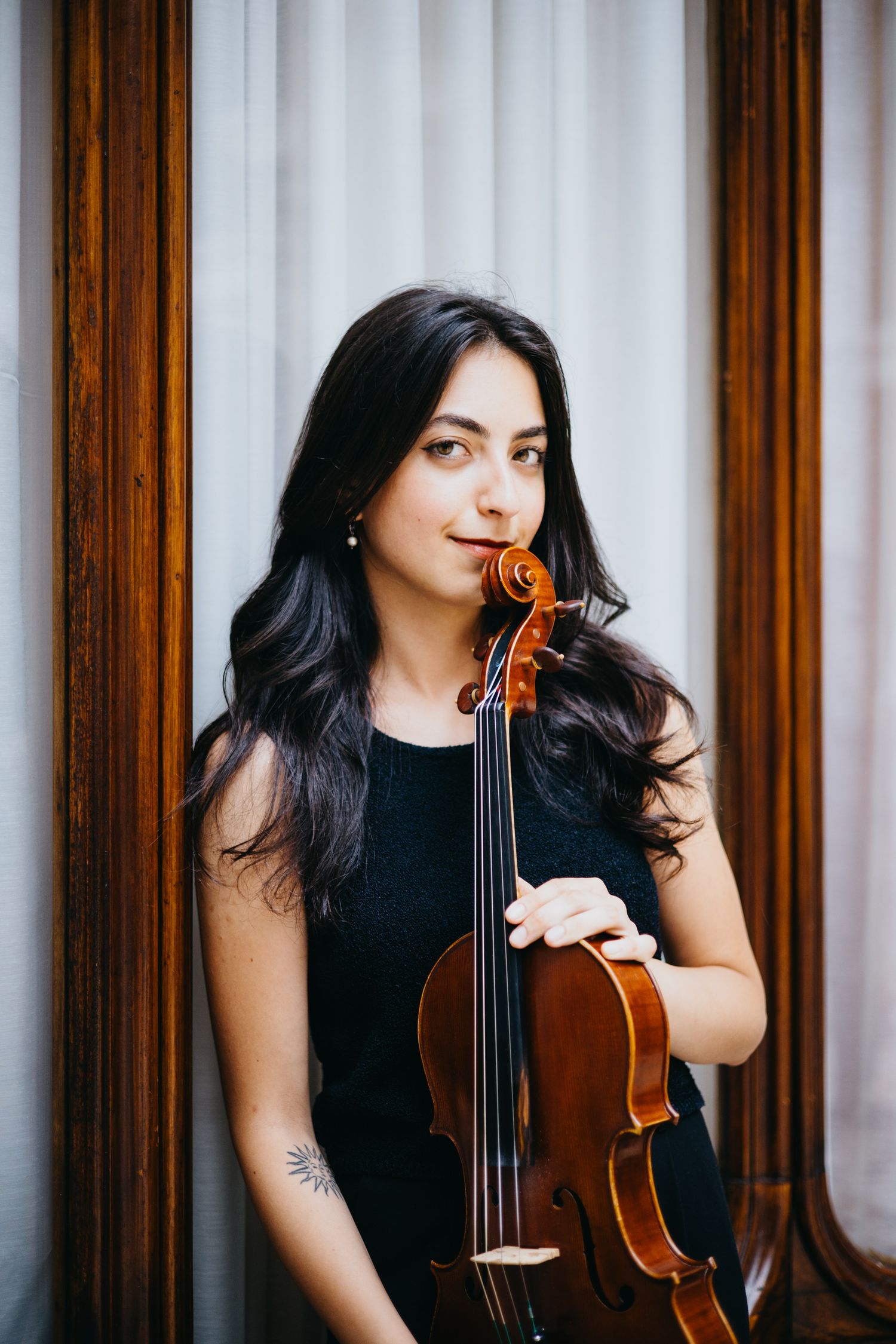 Dark-haired female violinist with tattoo holds violin near wooden framed window indoors.