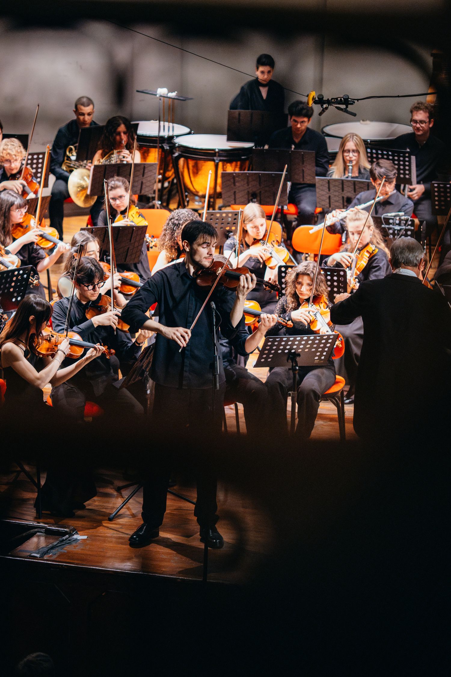 A male violinist performs as a soloist in a black outfit before a large orchestra on a concert stage.