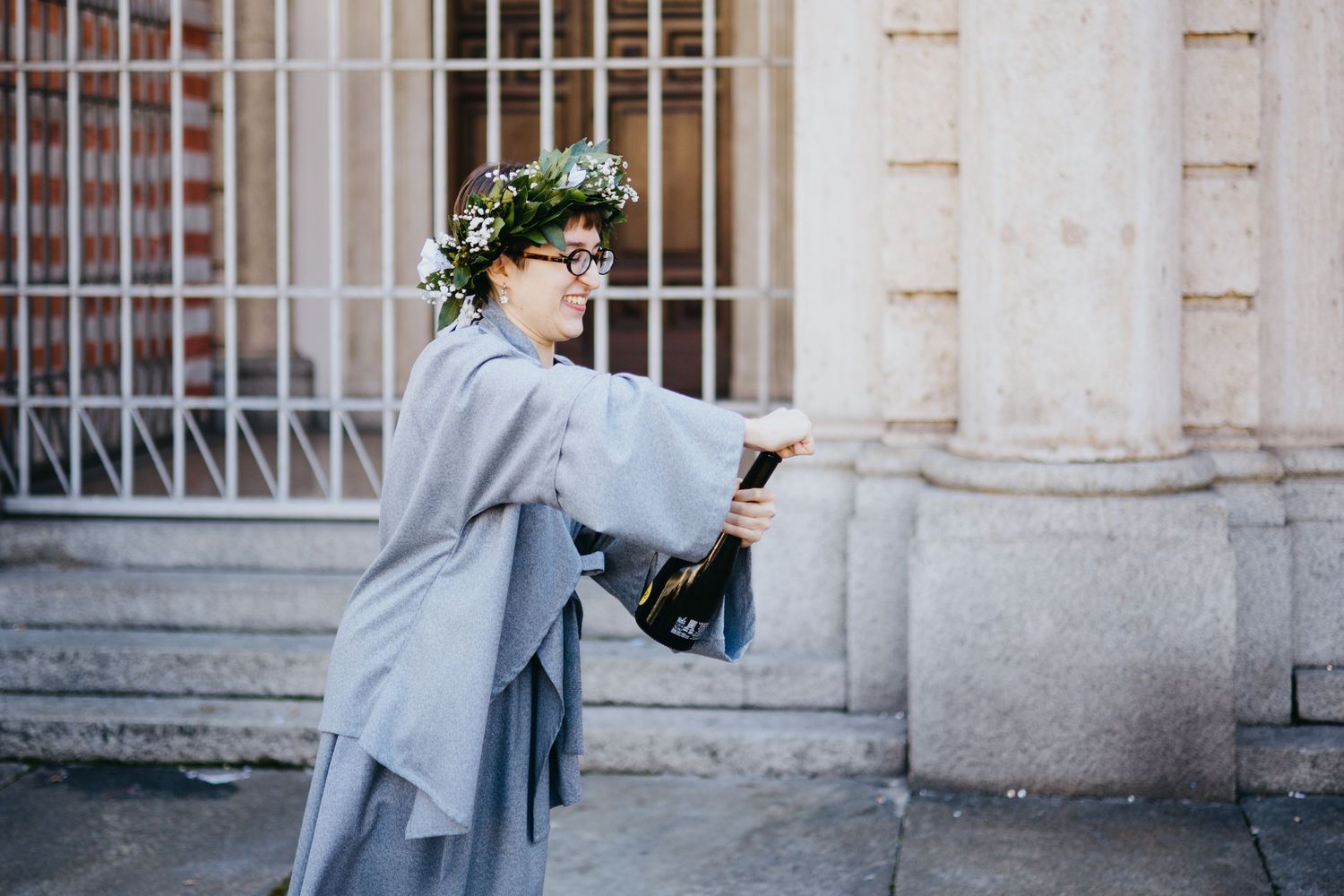Woman in gray graduation robe with floral crown pops champagne bottle outside building.