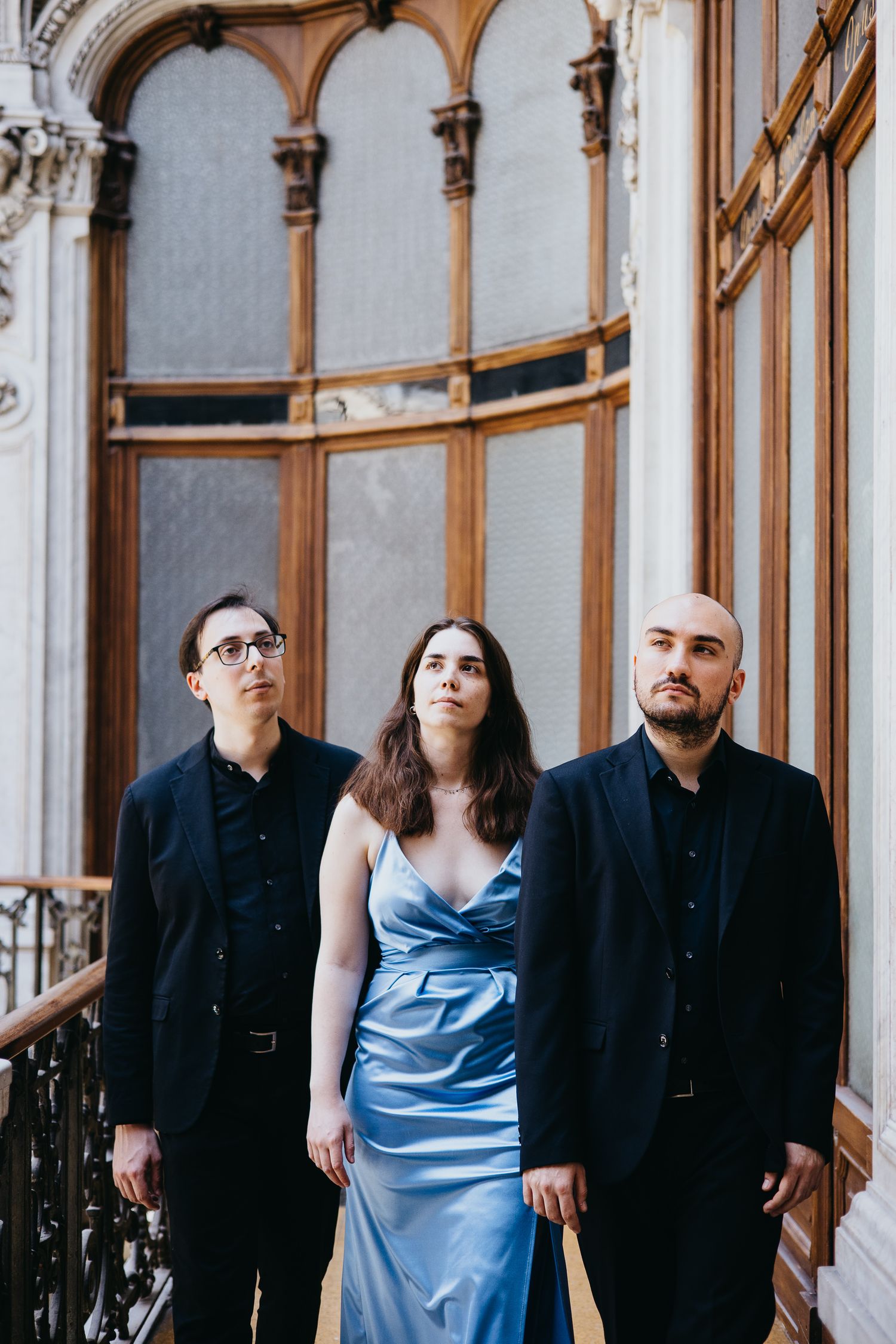 Three classical musicians in formal attire pose solemnly before ornate wooden doors on a balcony.