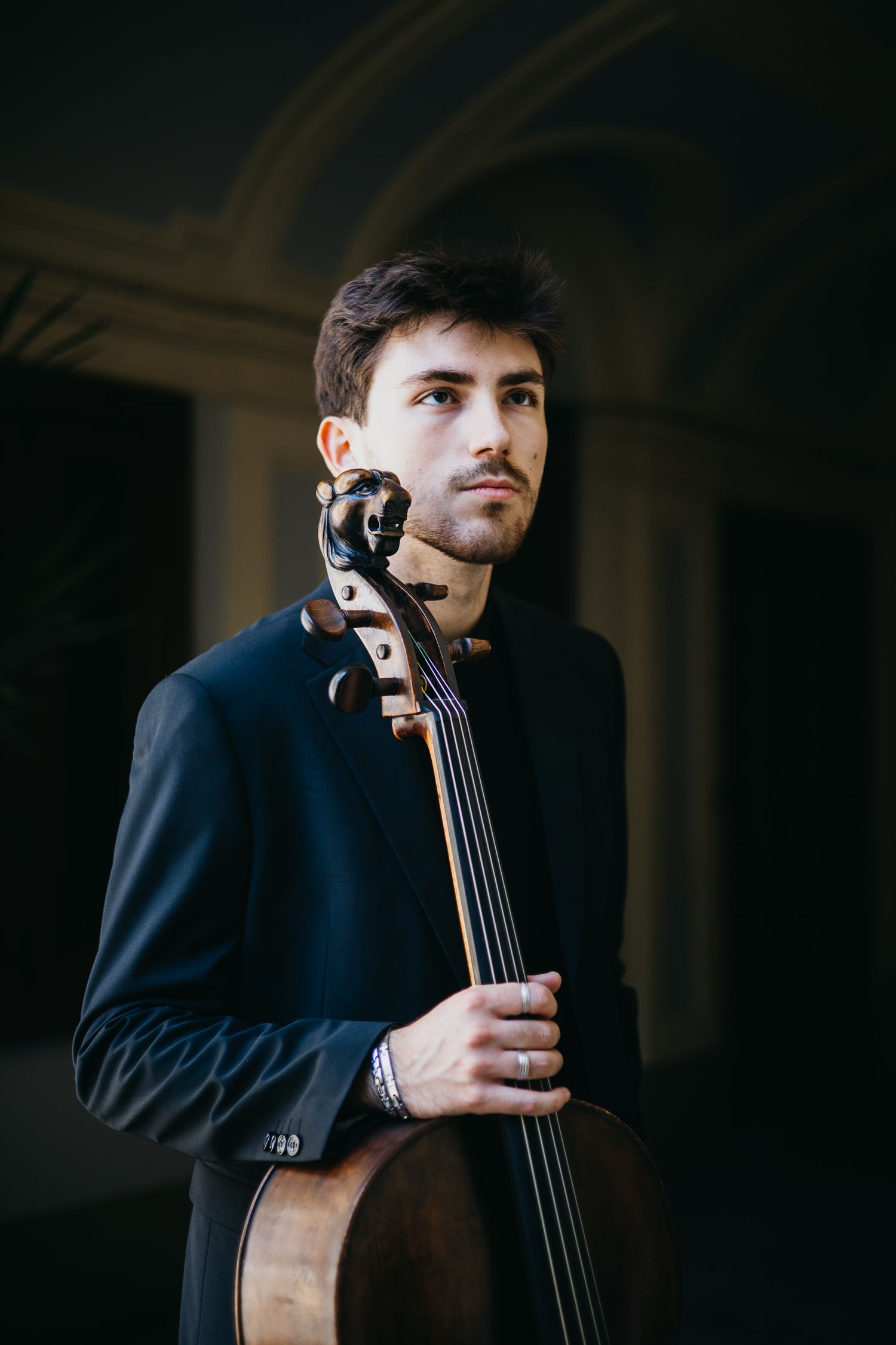Male cellist in black suit holding cello in moody dark architectural interior, looking away.