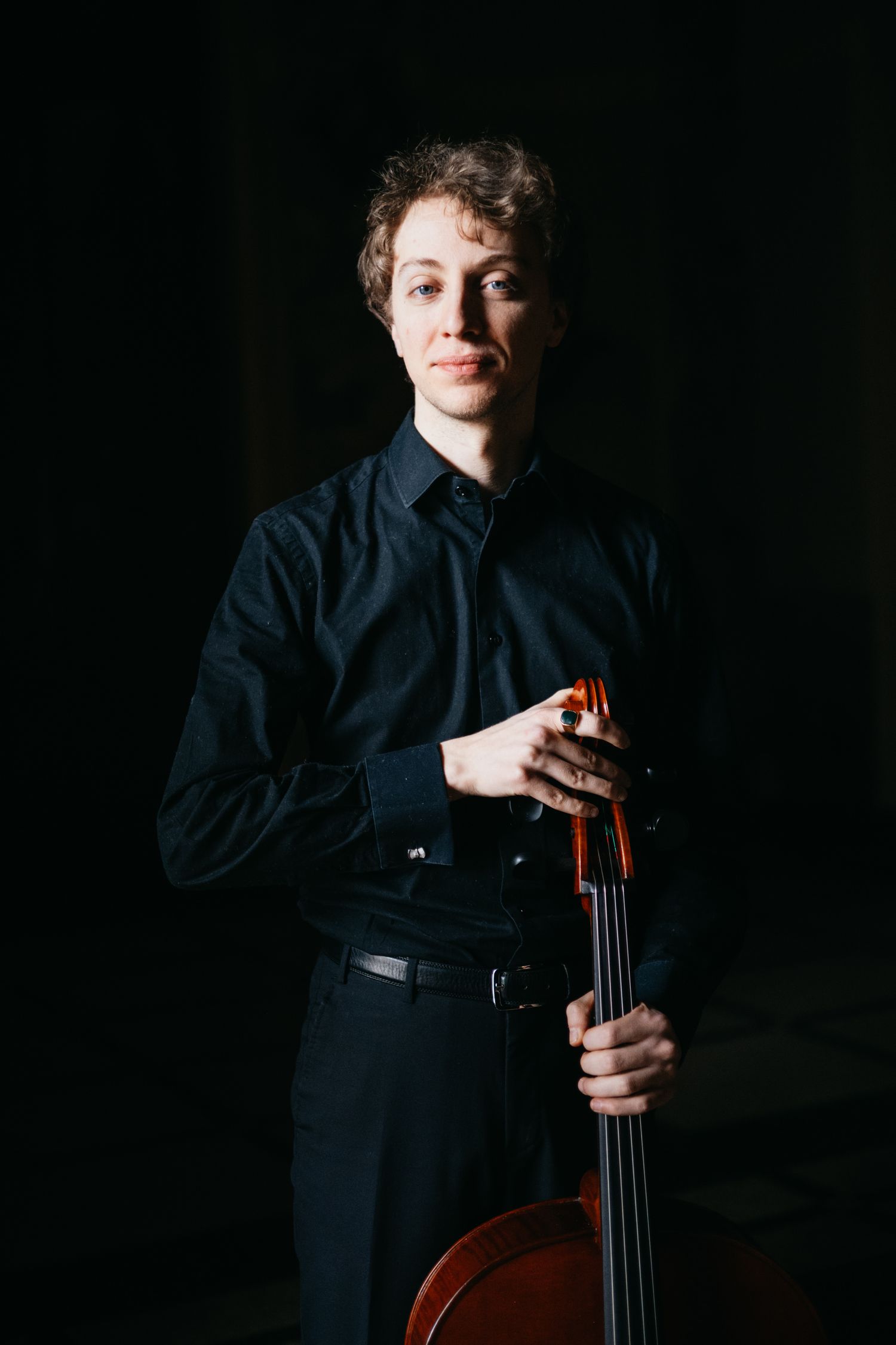 Young cellist in black attire poses with cello in dramatic dark lighting.