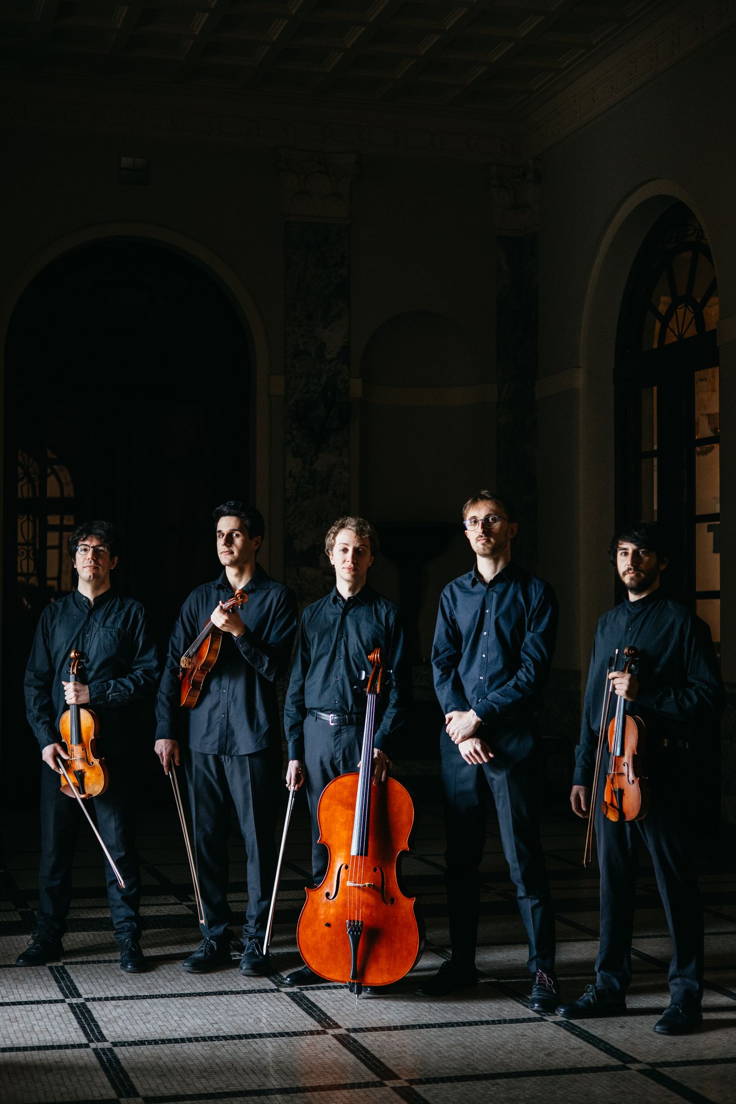 Five string musicians in black holding violins and cello pose in dramatic arched hallway.