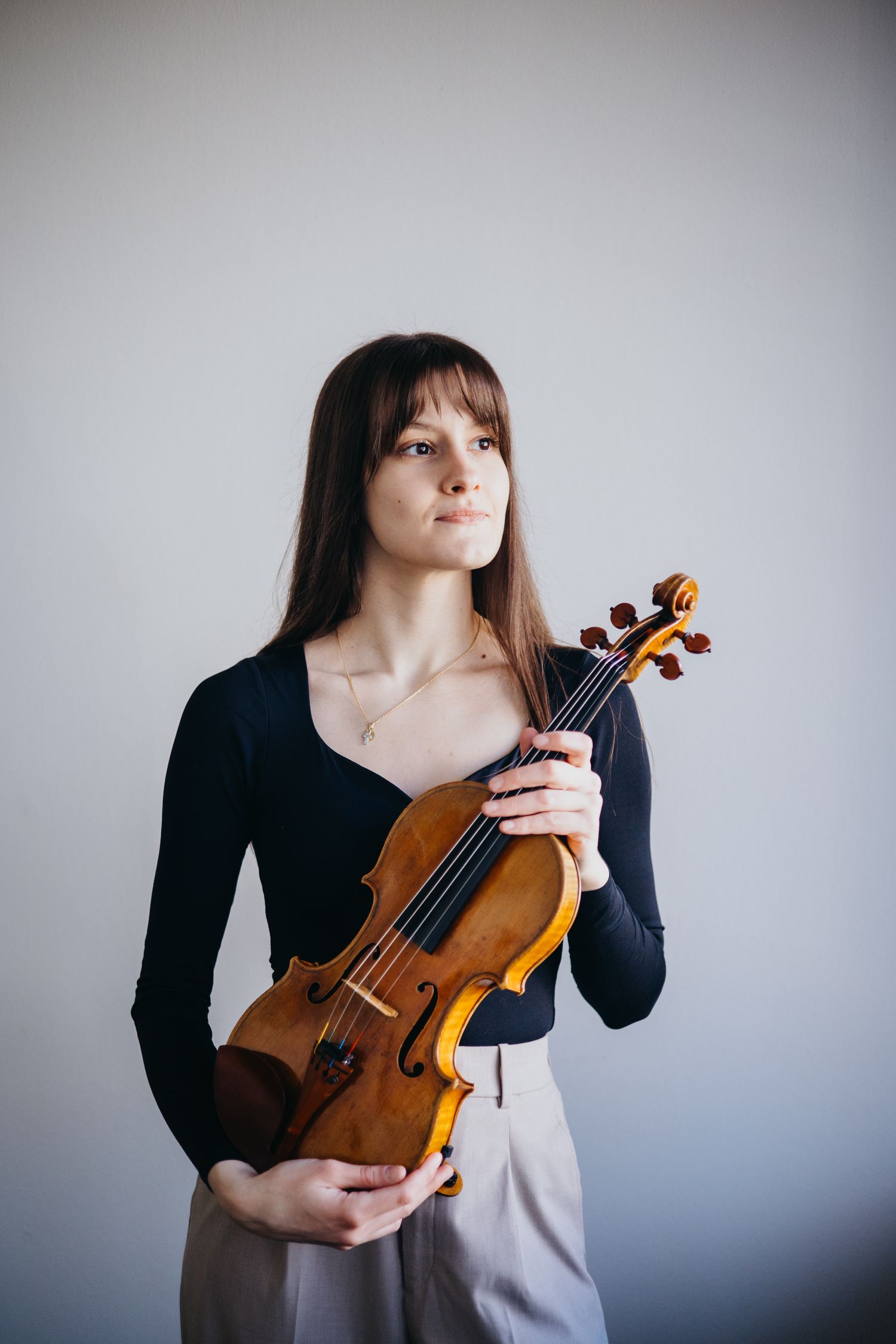 Female musician in black top and beige trousers holding a violin against white wall.