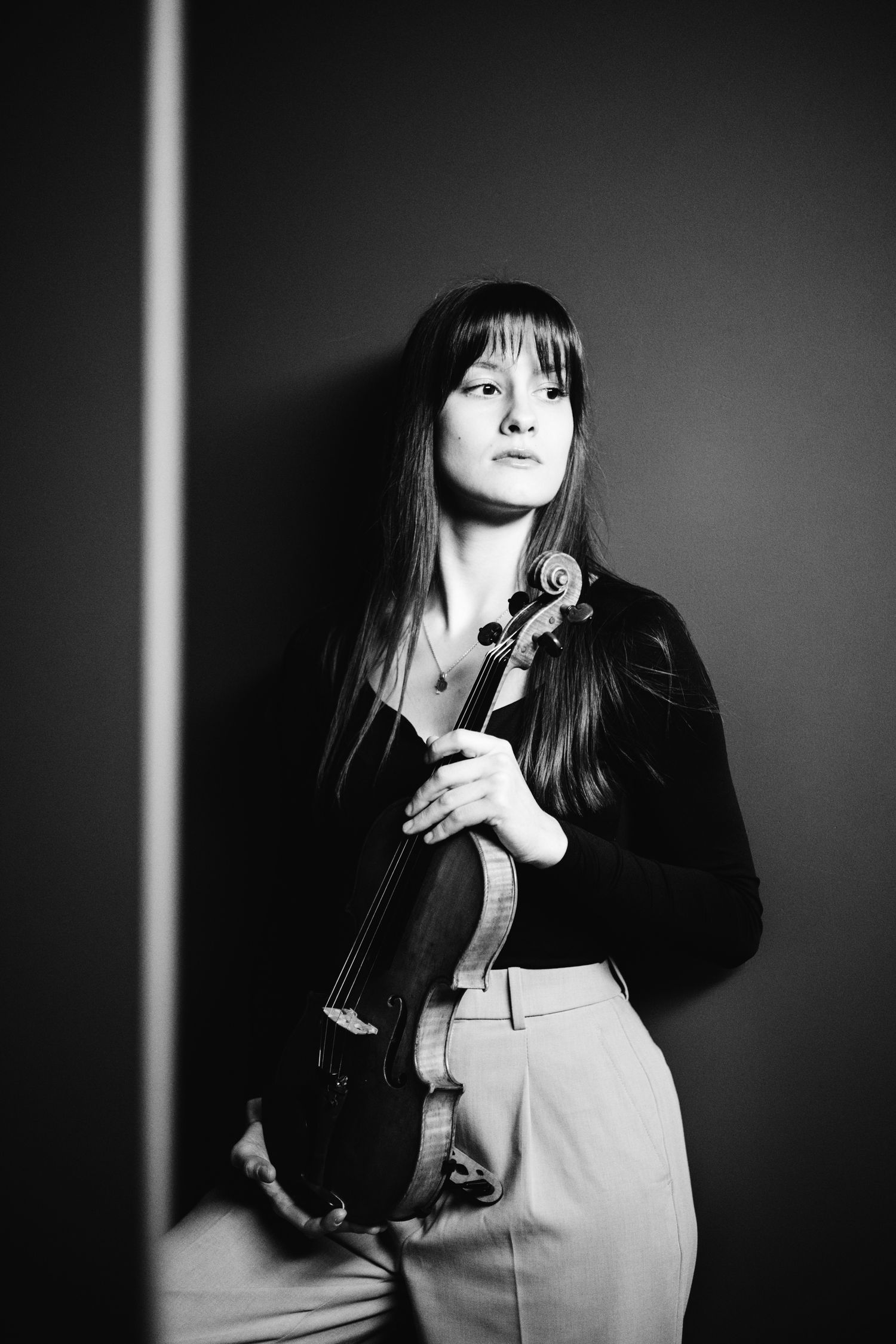 Young female violinist holds her violin while leaning against a wall in a black and white portrait photo.