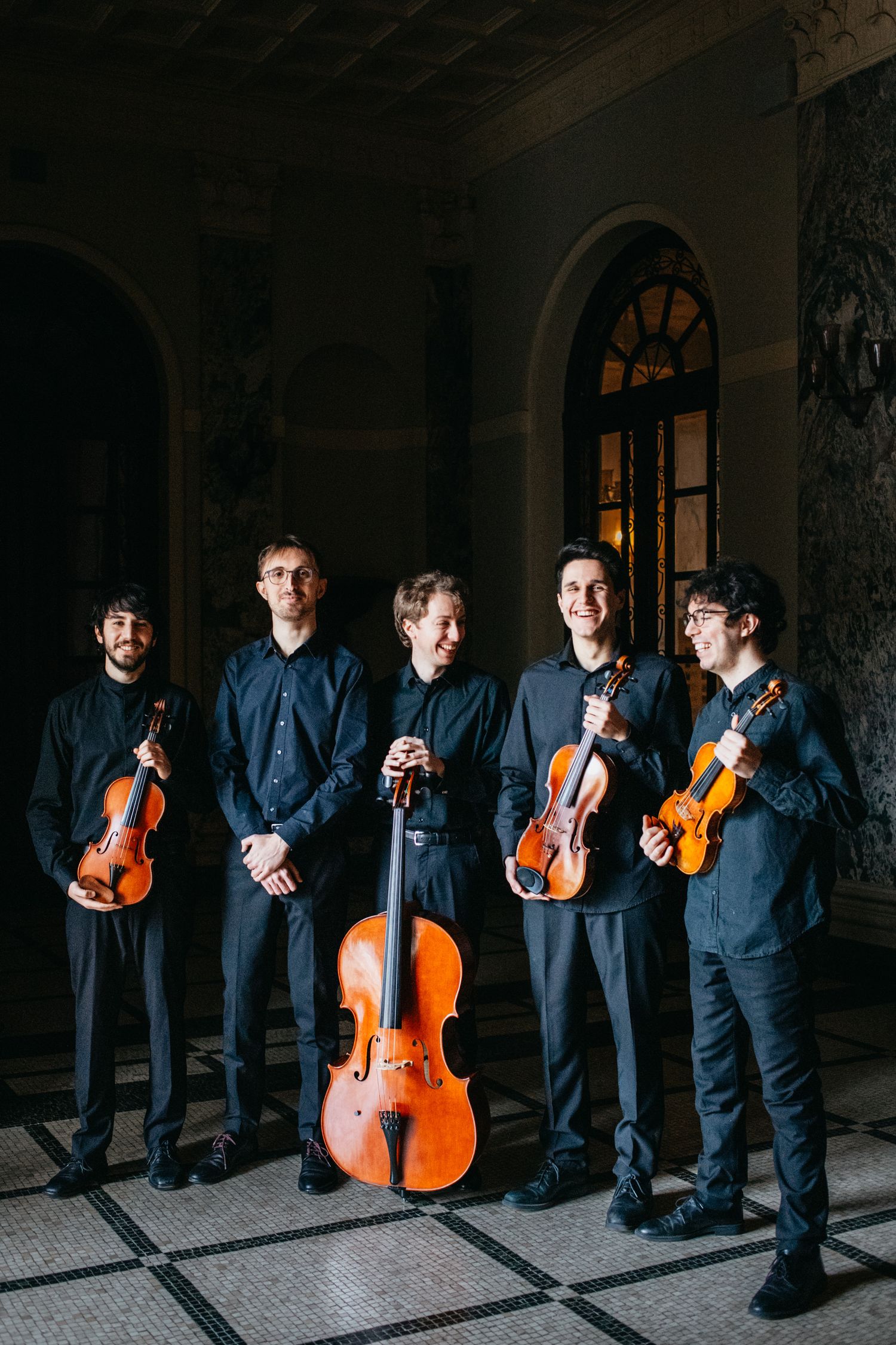 Five male string musicians smiling and holding instruments in ornate classical interior.