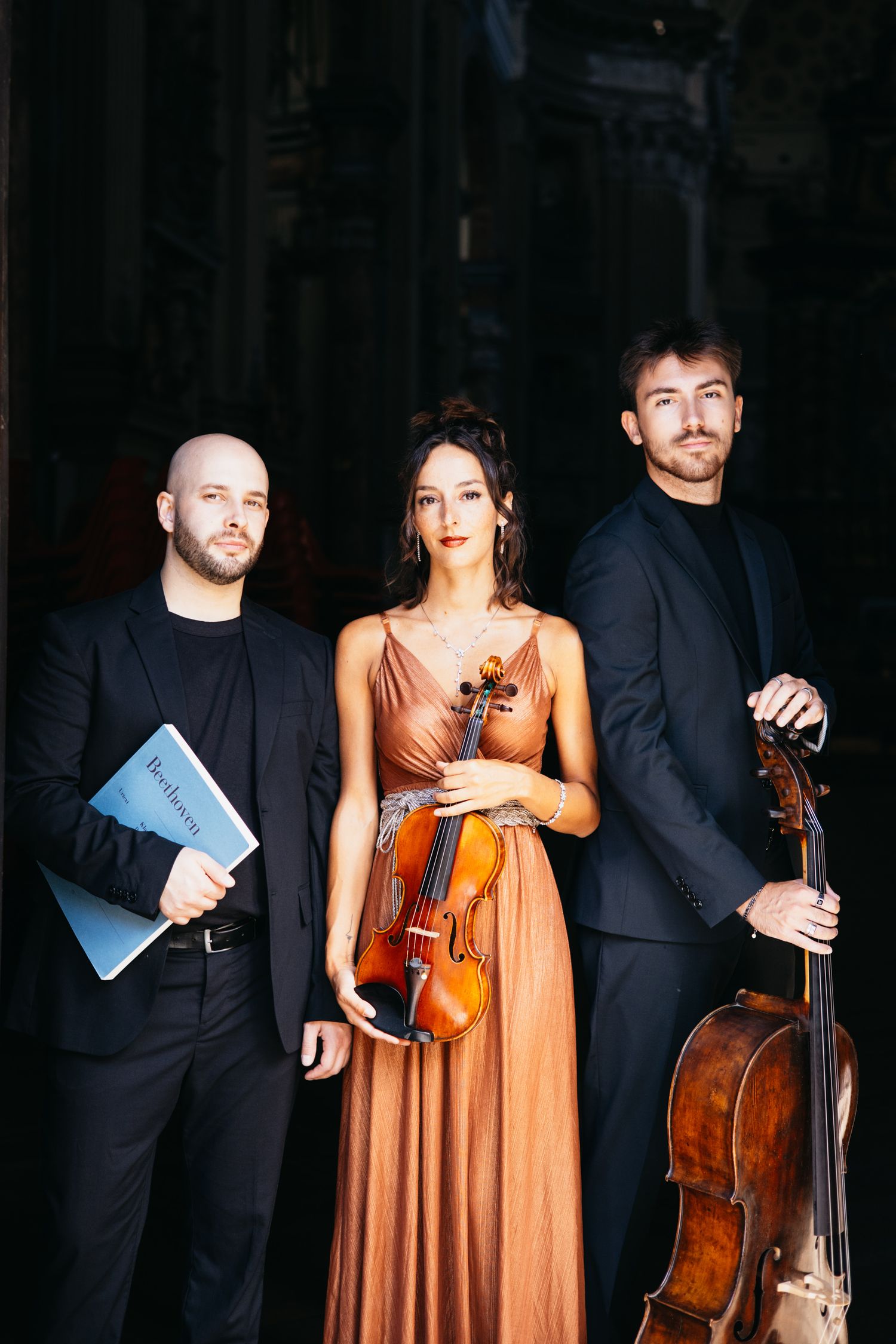 Three classical musicians in formal attire pose with violin and cello instruments in an elegant dark setting.