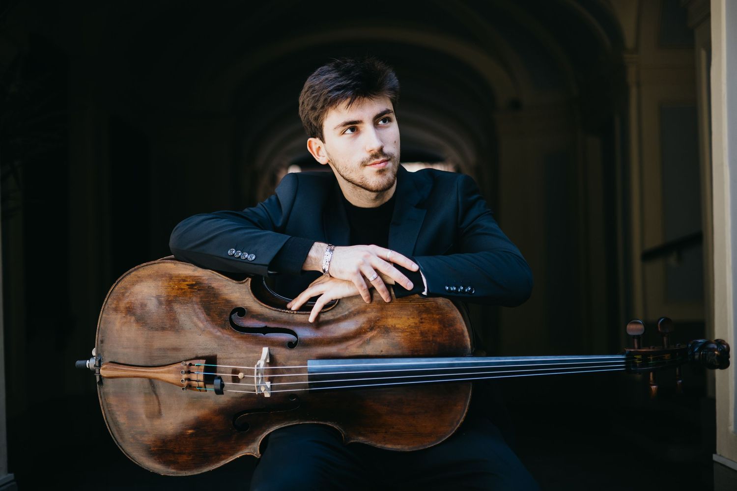 Young male cellist leaning on cello while seated in classical stone archway setting.