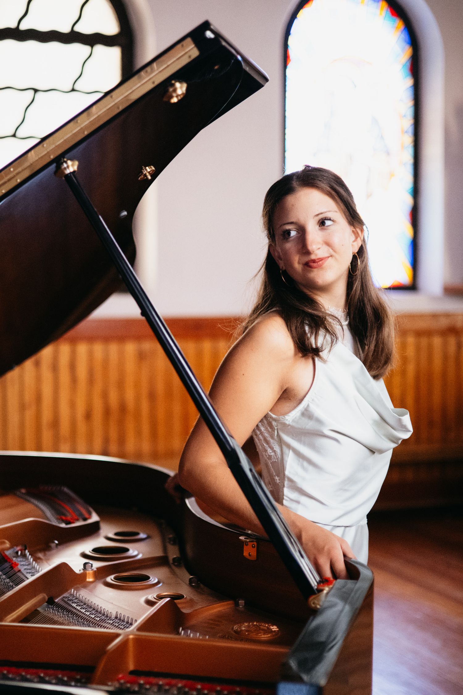 Young woman in white dress smiling beside an open grand piano in a church with stained glass windows.