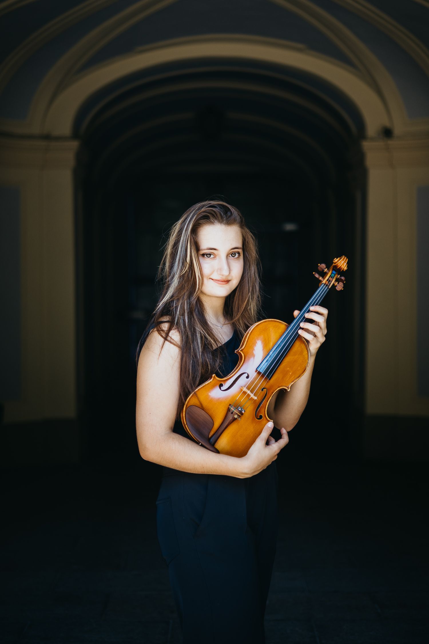 Young female violinist in black sleeveless dress poses with violin under stone arch.