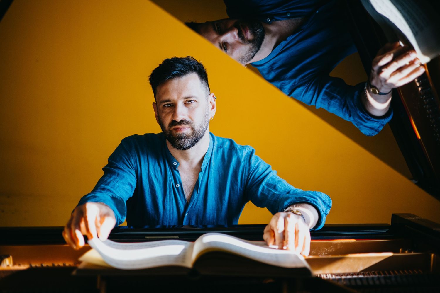 Male pianist in blue shirt reviewing sheet music at grand piano with yellow background.