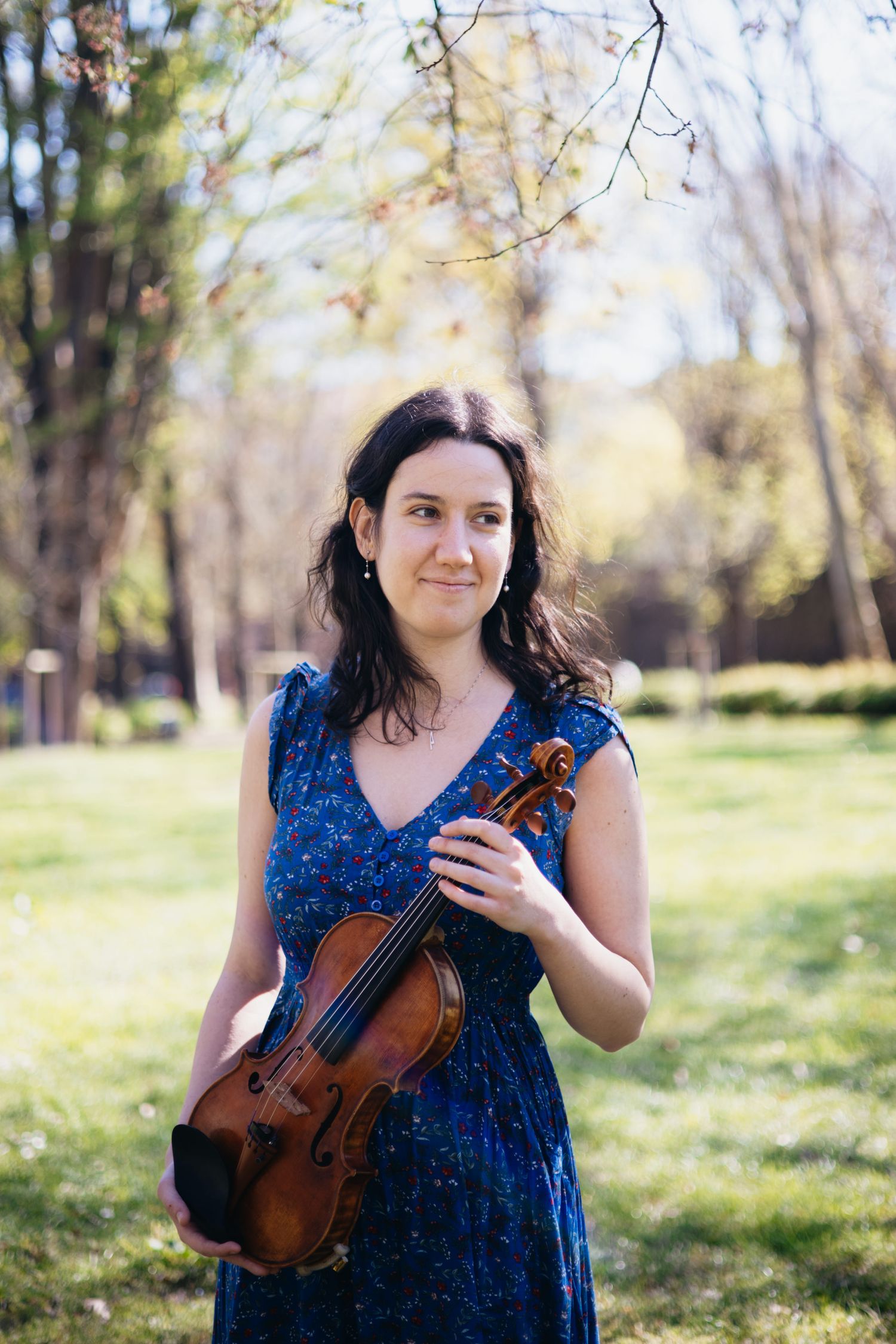 Female violinist in blue floral dress holding violin outdoors in sunny green park.