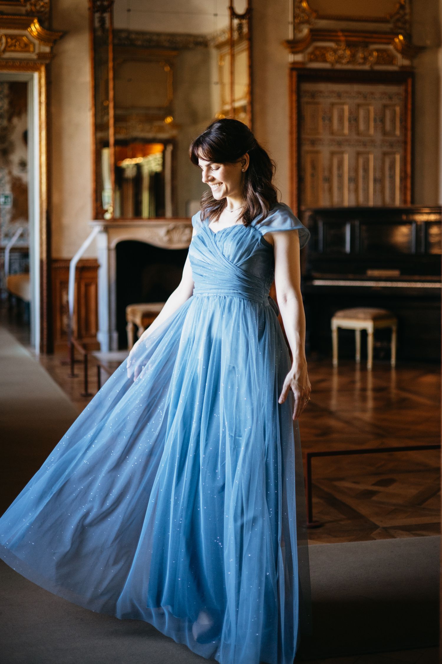 Woman in flowing blue gown twirling in ornate ballroom with fireplace and piano.