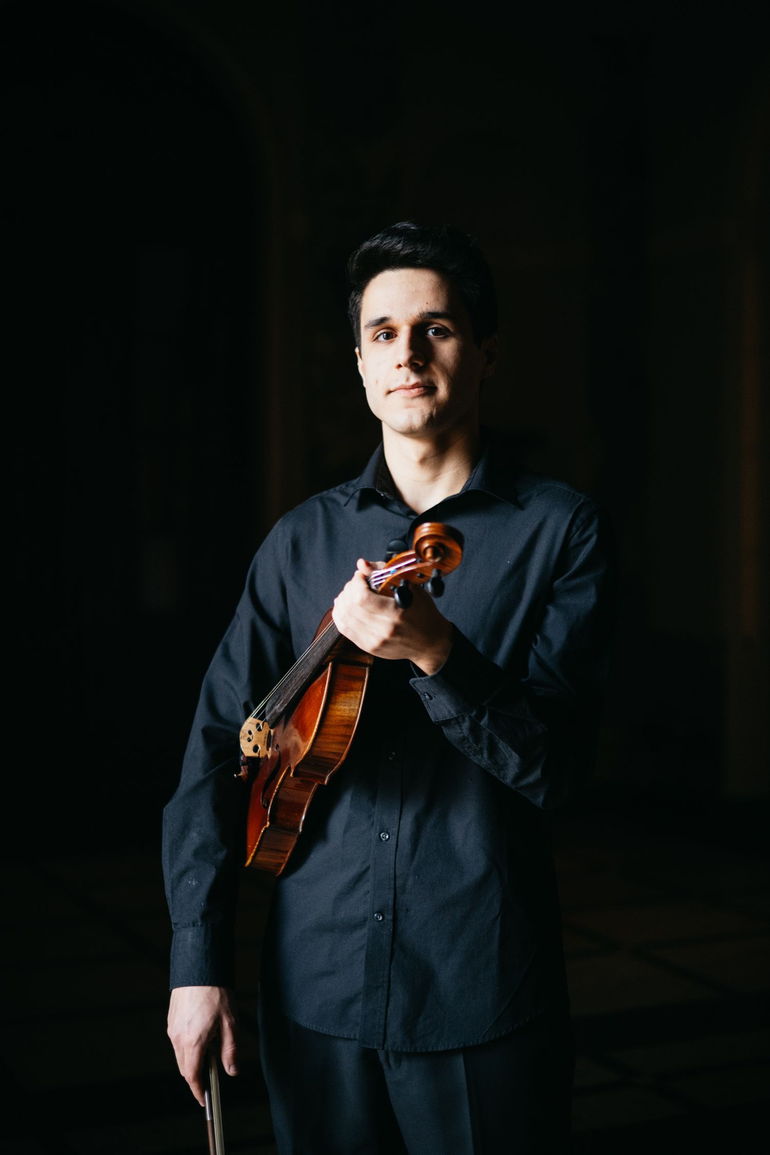 Male violinist in black clutches violin to chest in moody dark portrait photography.