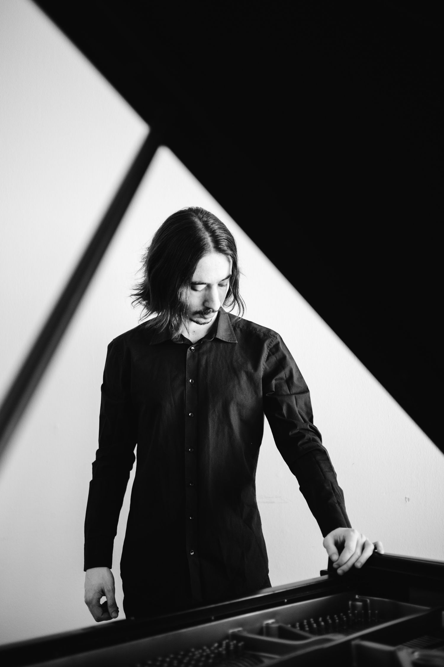Black and white portrait of a long-haired musician standing at a grand piano keys.
