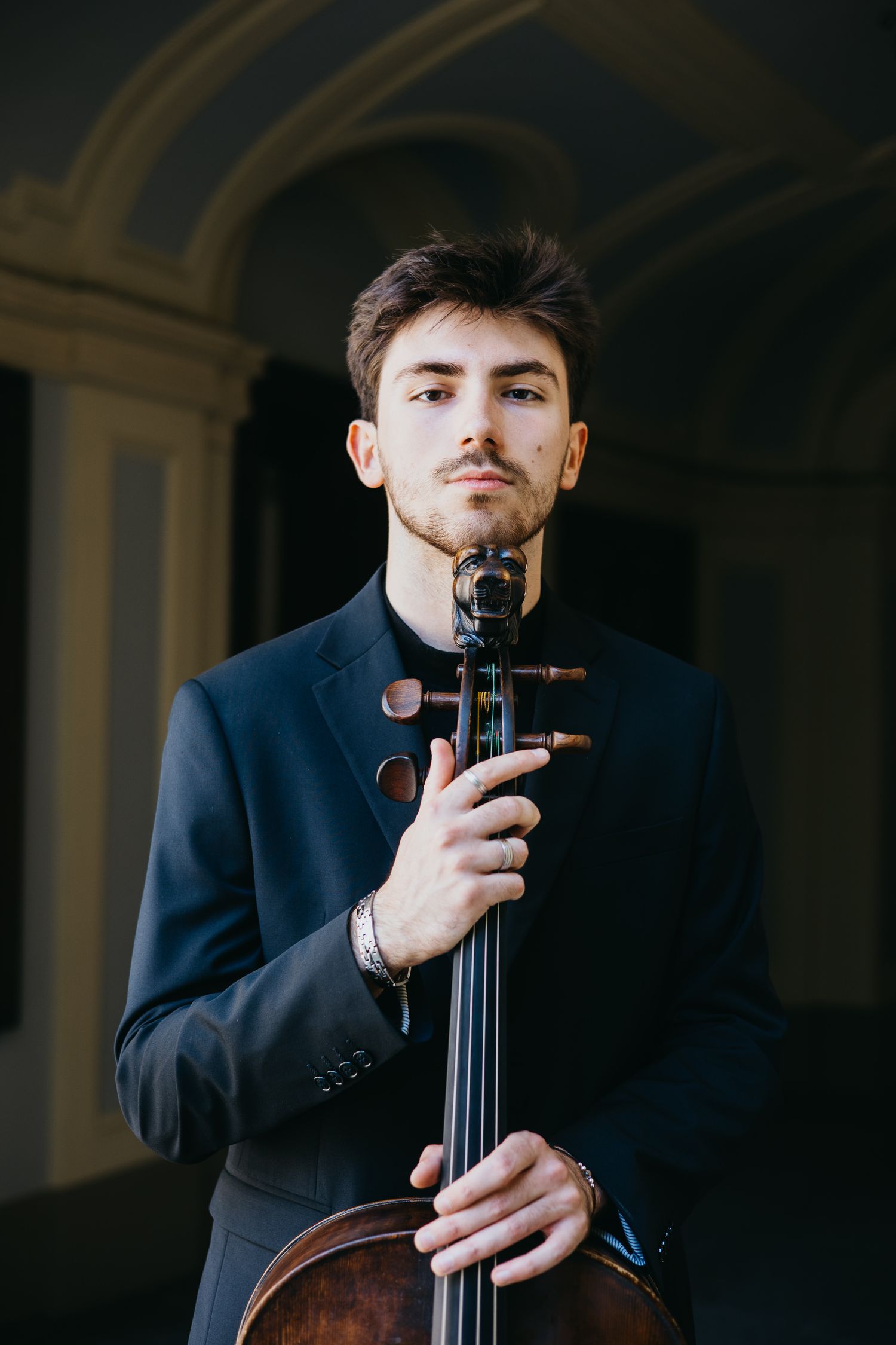 Young male classical musician holding cello upright in dark architectural setting wearing black jacket.