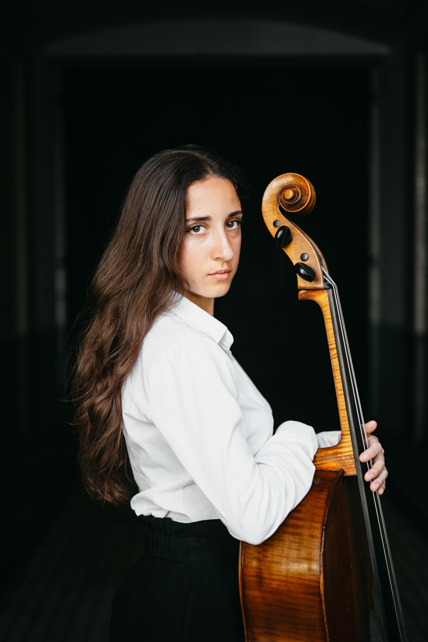 Young female cellist in white shirt poses with cello against a dark background.