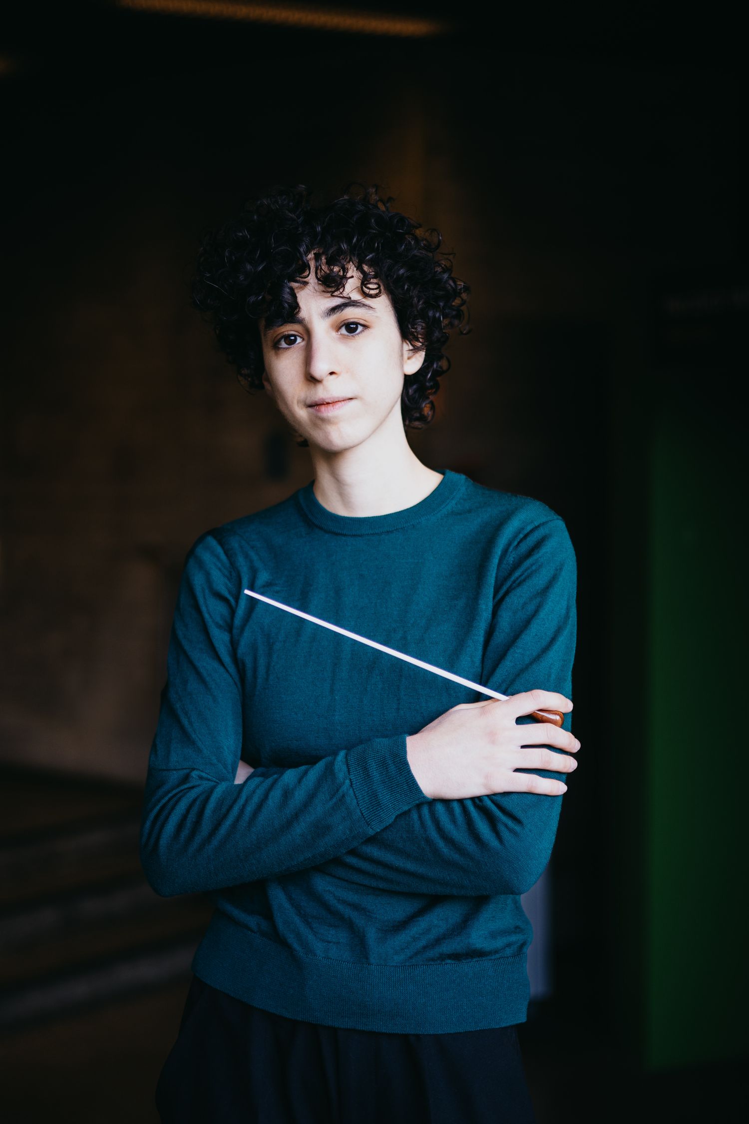 Young conductor with curly hair holding baton with arms crossed.