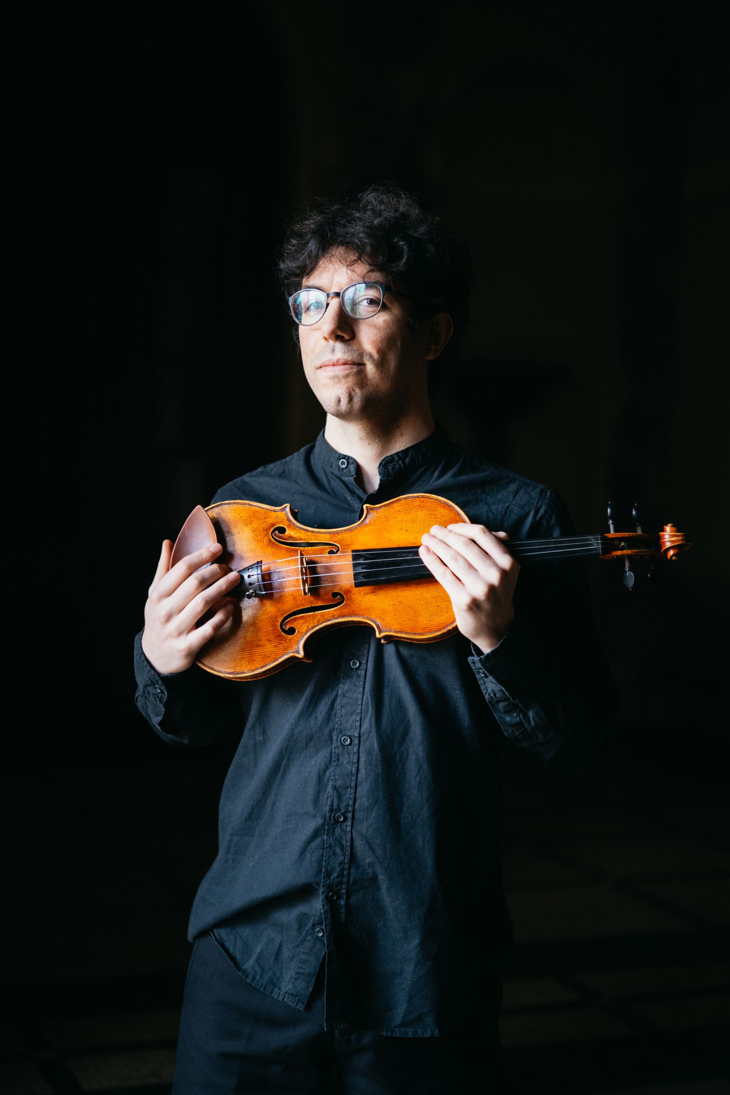 Male violinist wearing glasses holding violin in dark dramatic studio portrait setting.