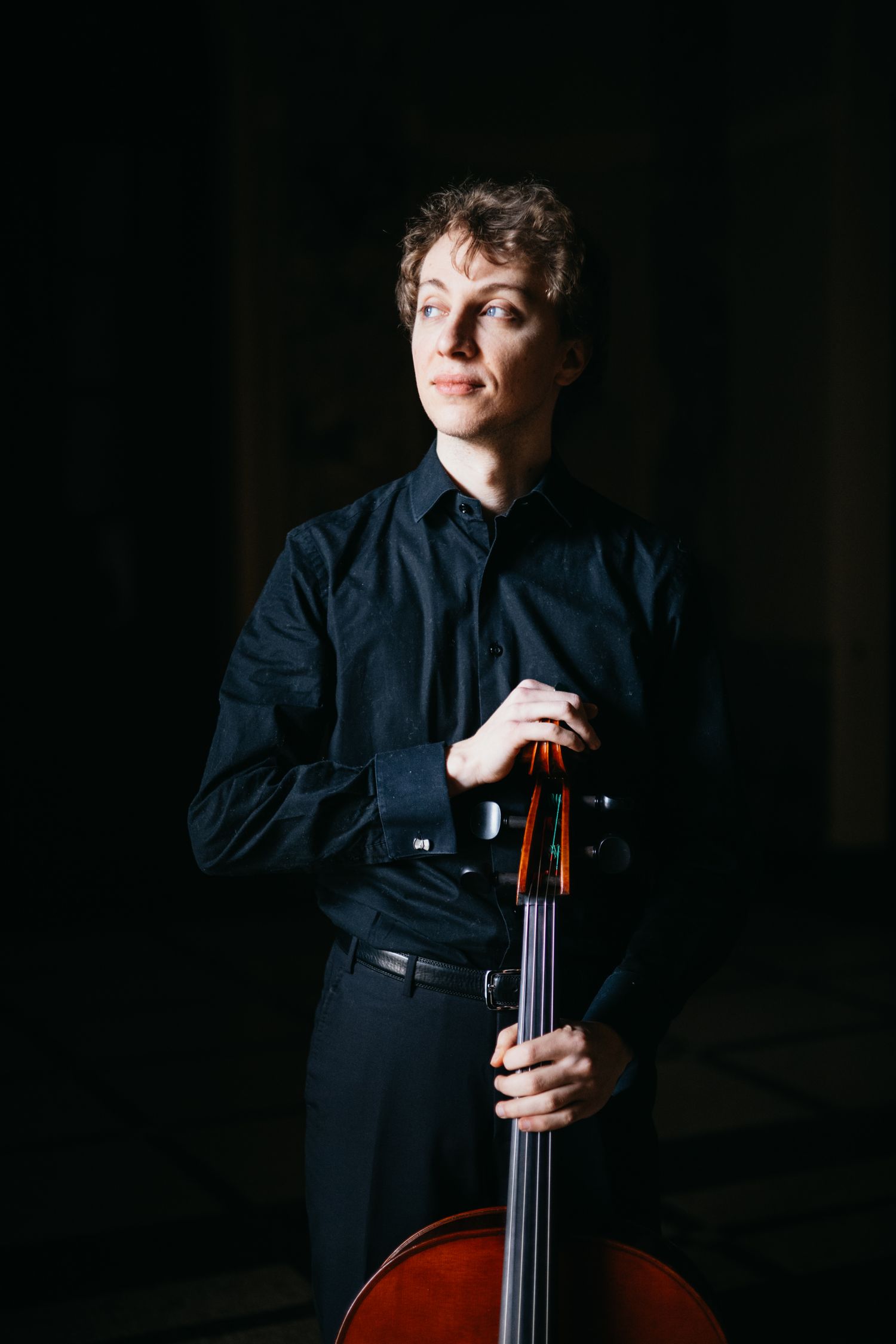 Young cellist in black shirt rests hands on cello scroll in dark setting.