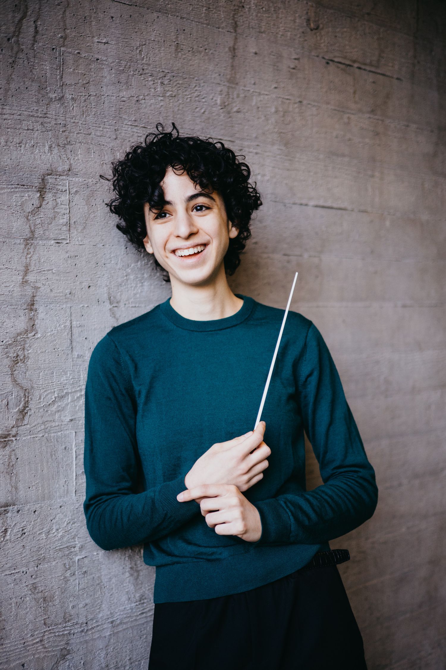 Smiling young male conductor with curly hair holds a conductor baton against a concrete wall.