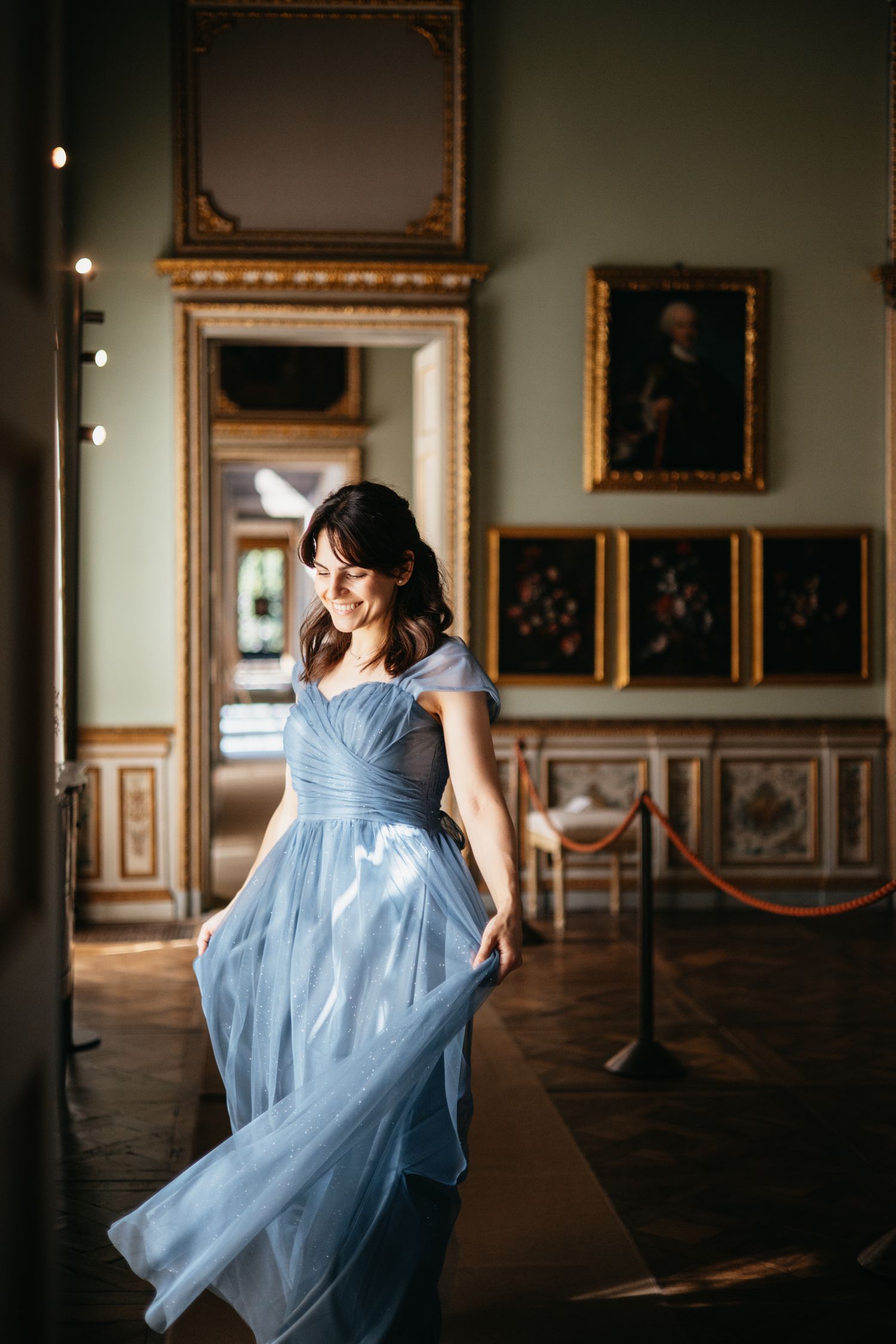 A woman in a flowing light blue gown twirls gracefully in an elegant gallery room with ornate gold-framed paintings.
