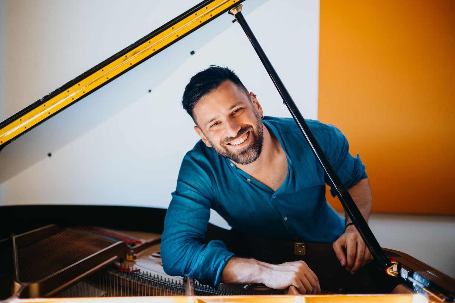 Smiling bearded pianist in blue shirt leaning on grand piano in a bright colorful room.