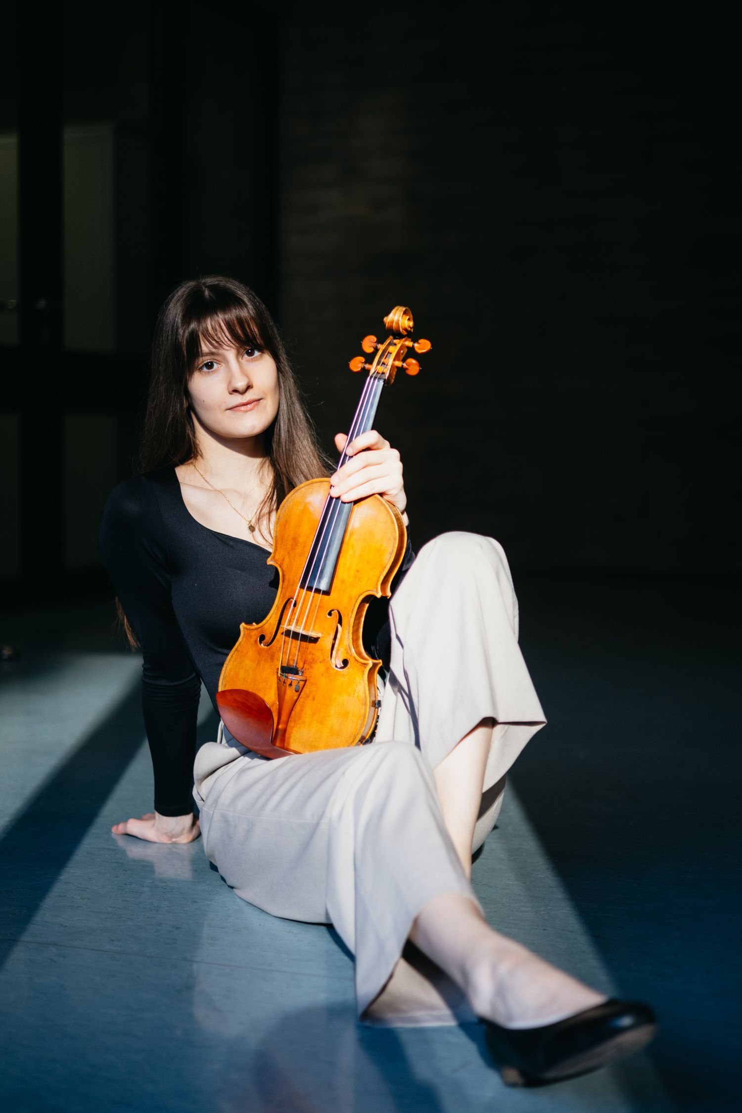 Young woman sits on floor holding a violin upright in dramatic chiaroscuro lighting.