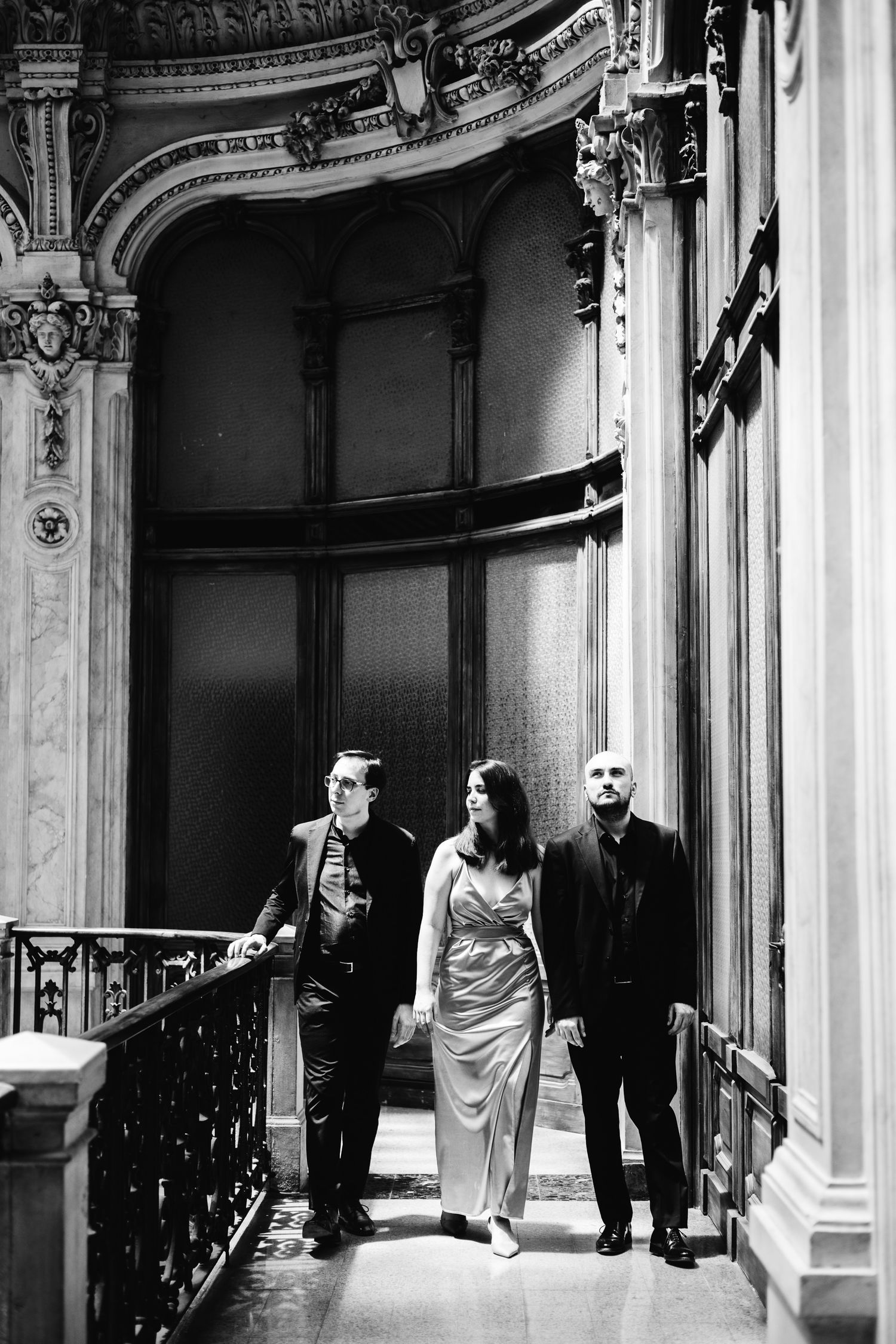 Three classical musicians in formal attire walk through an ornate grand building interior in black and white.