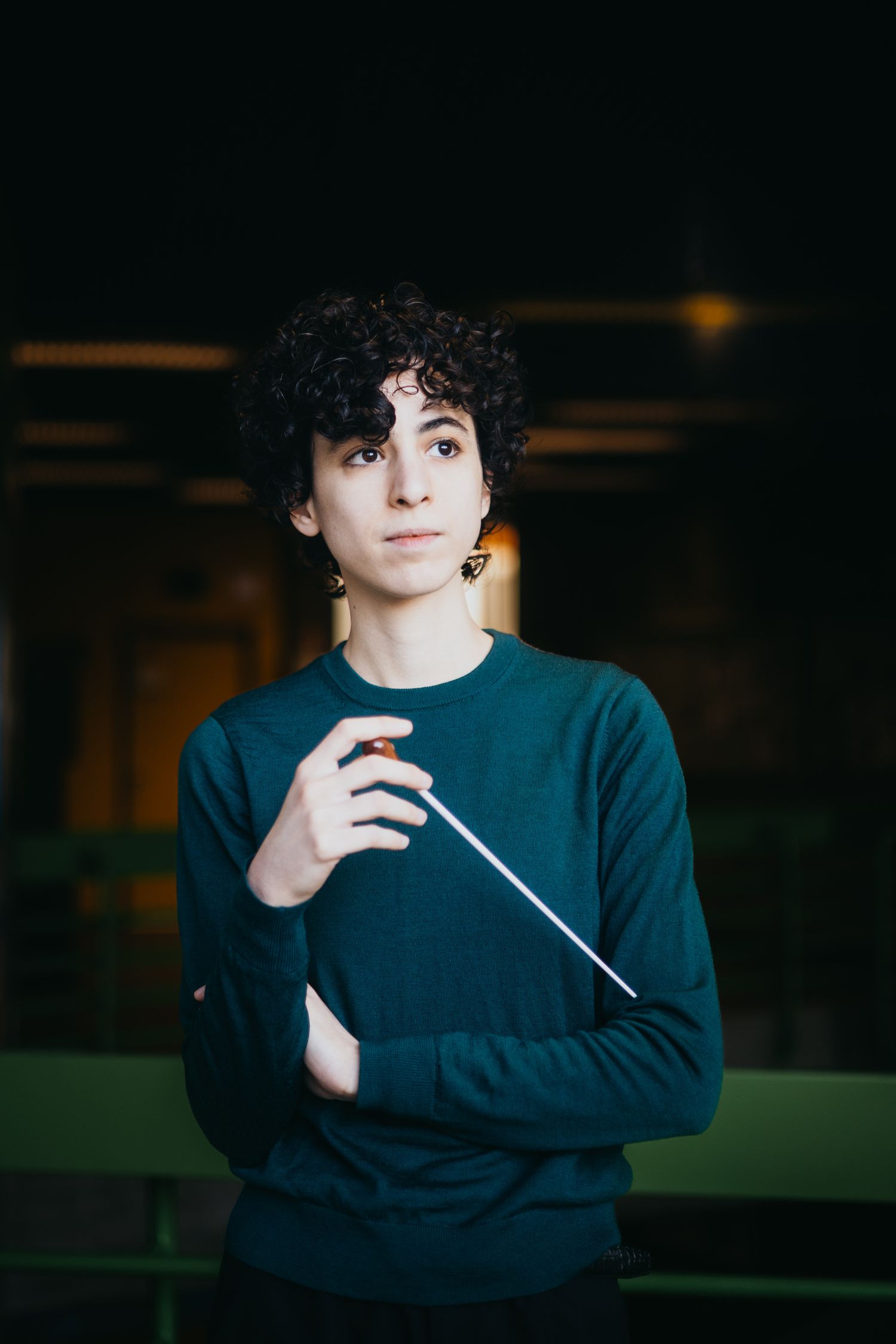 Young conductor in teal sweater holding baton in dark interior setting.