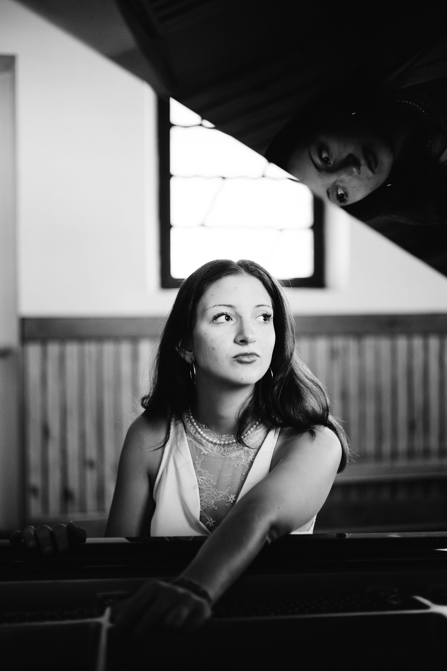 Black and white portrait of a young woman in pearls sitting at a grand piano, gazing upward thoughtfully.