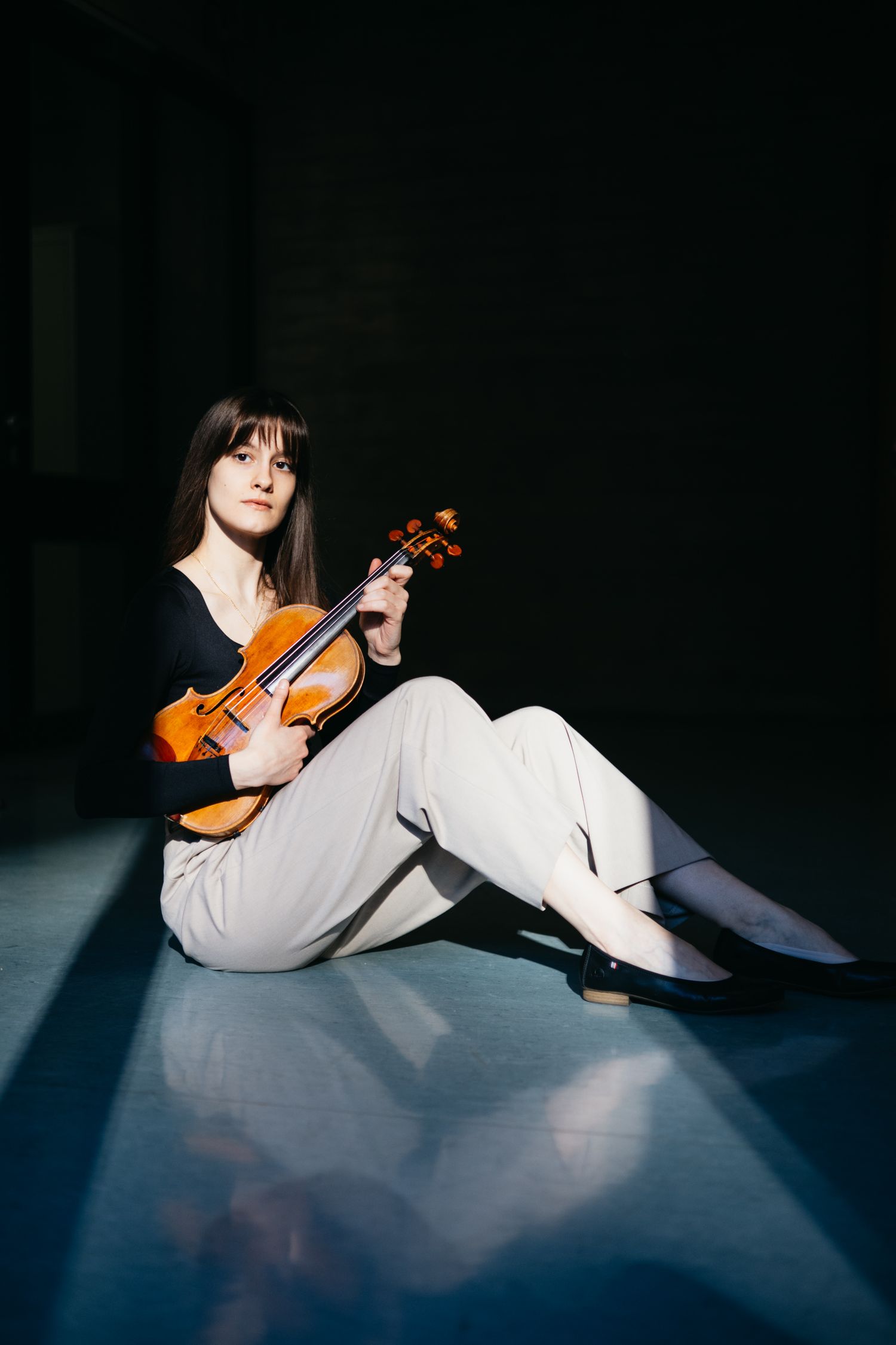 Female violinist sits on the floor cradling her violin in dramatic spotlight lighting.