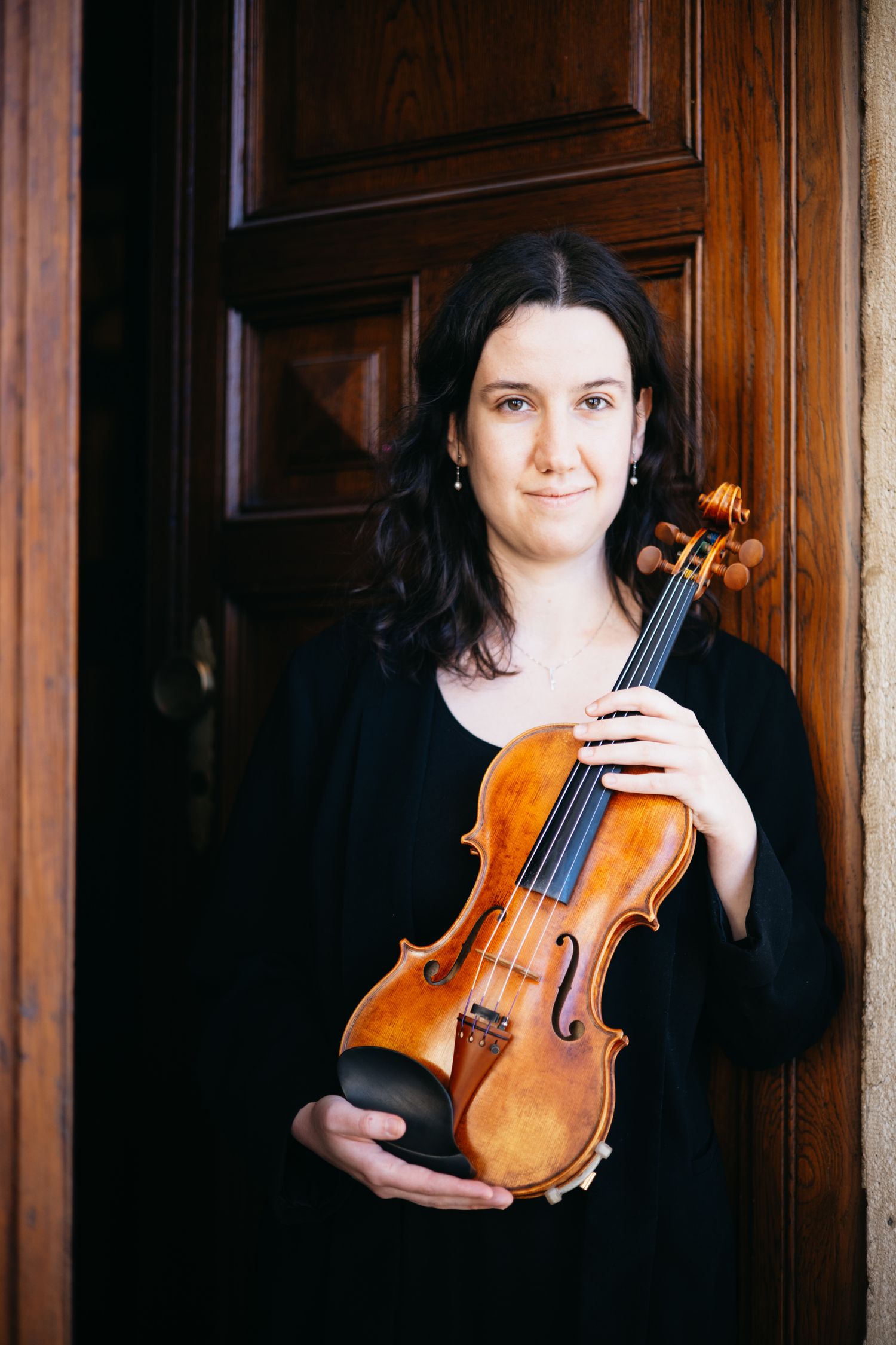 Woman in black dress holding a violin while standing in a wooden doorway, smiling at the camera.