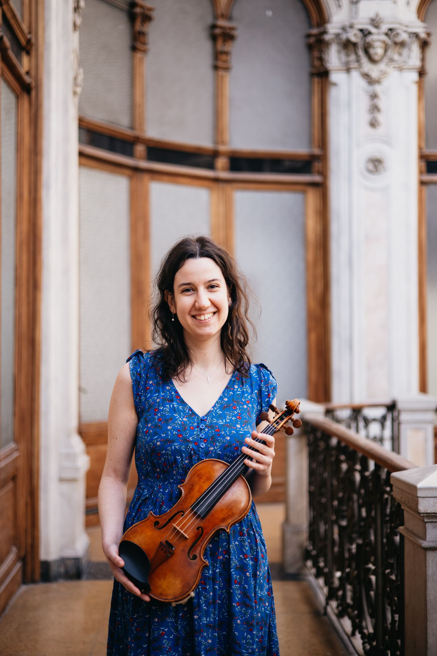 Smiling female violinist in blue floral dress holding violin in elegant architectural hallway.