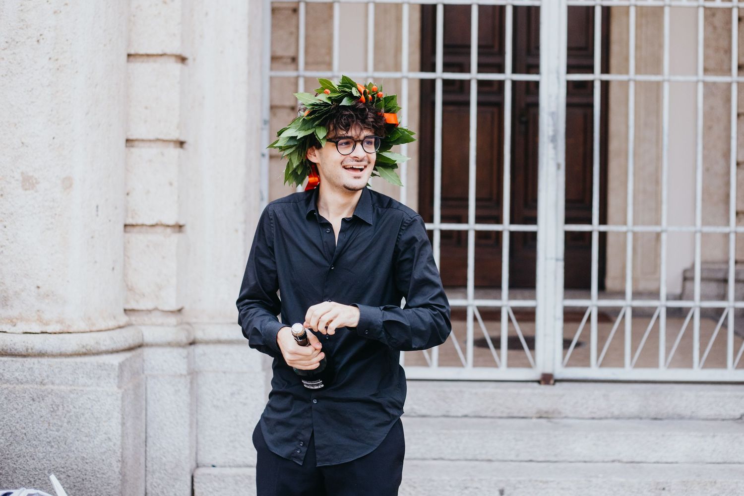 Smiling young man wearing a floral laurel wreath stands outdoors near a classical stone building.