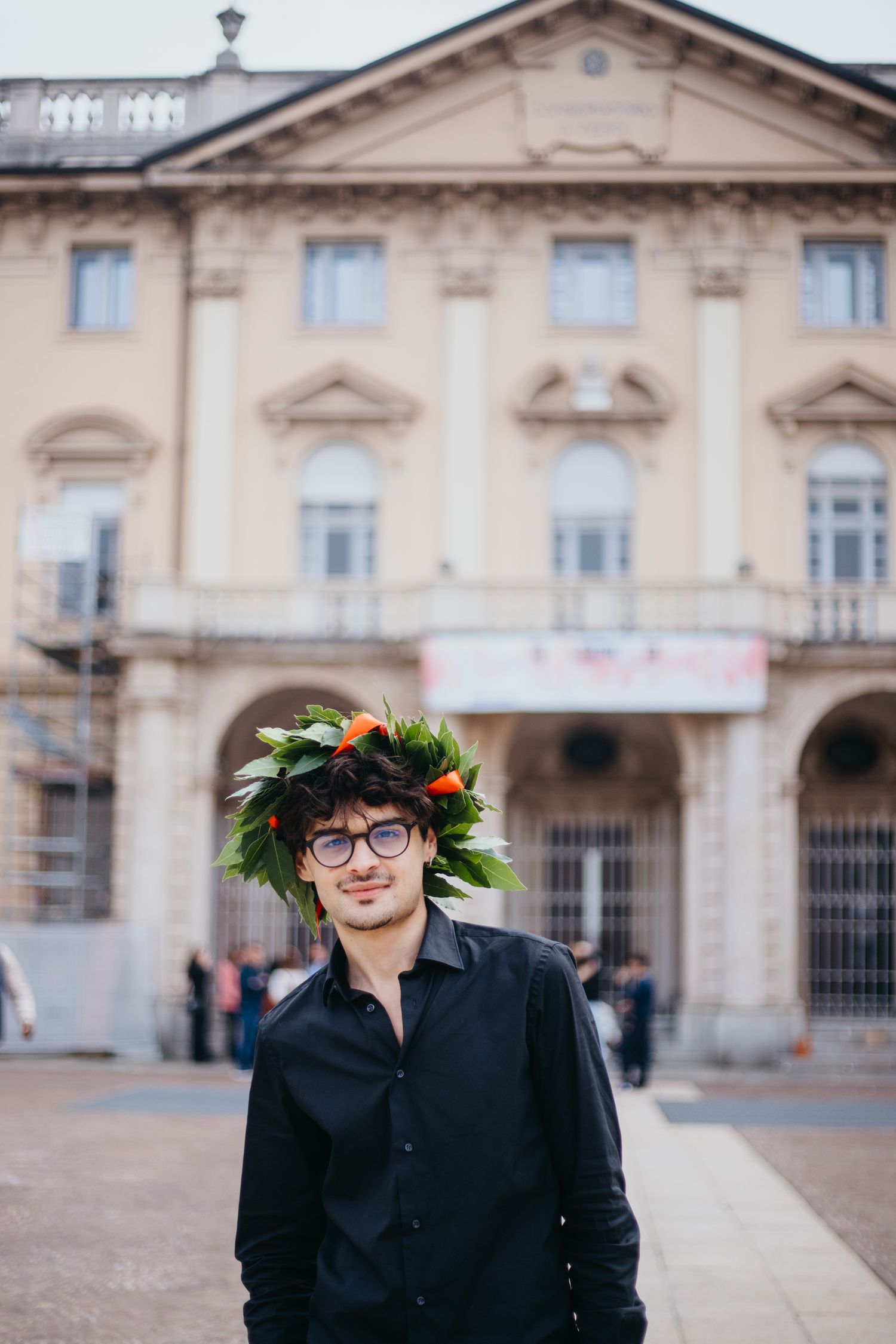 Young male graduate wearing laurel wreath stands smiling outside a classical Italian university.