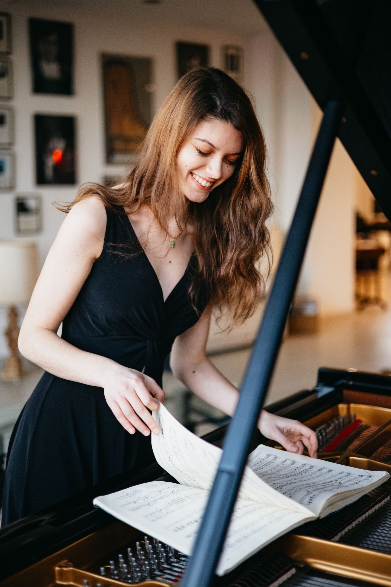 A smiling young woman in a black dress reviews sheet music at an open grand piano in an elegant room.