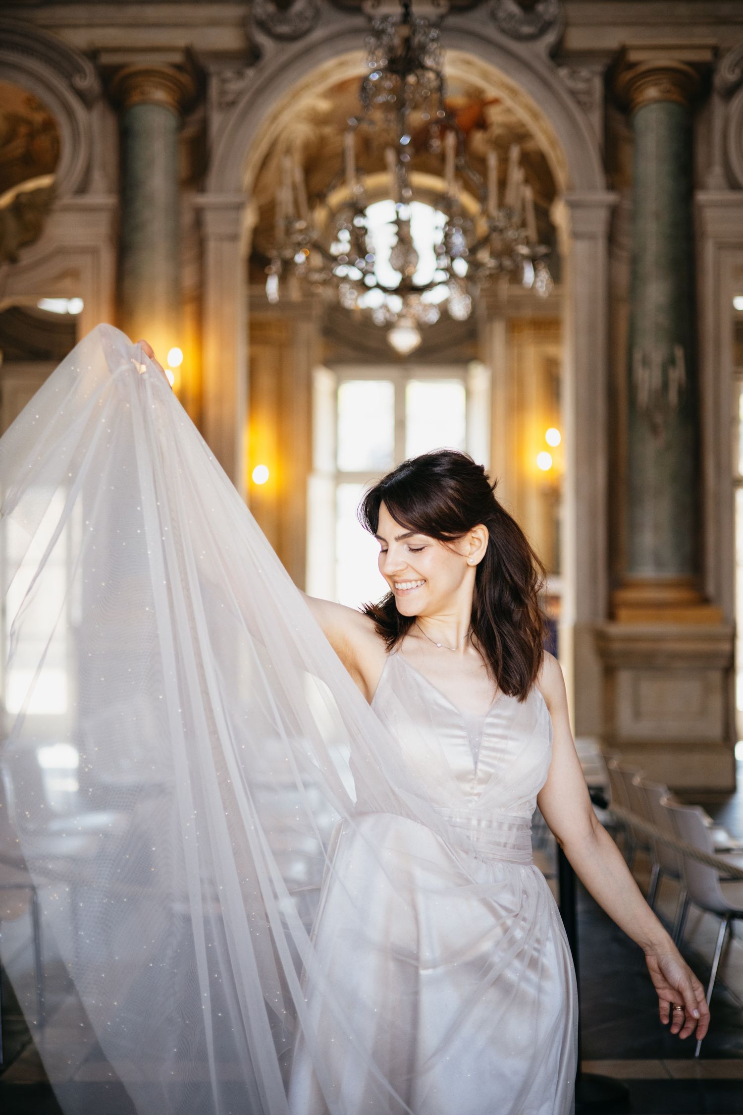 Smiling bride in white gown with flowing veil in ornate chandelier ballroom.
