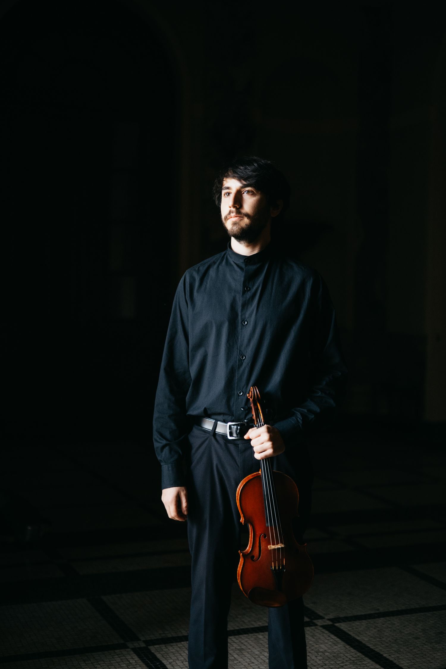 Young male violinist in black attire holding violin at side in dramatic dark studio portrait.