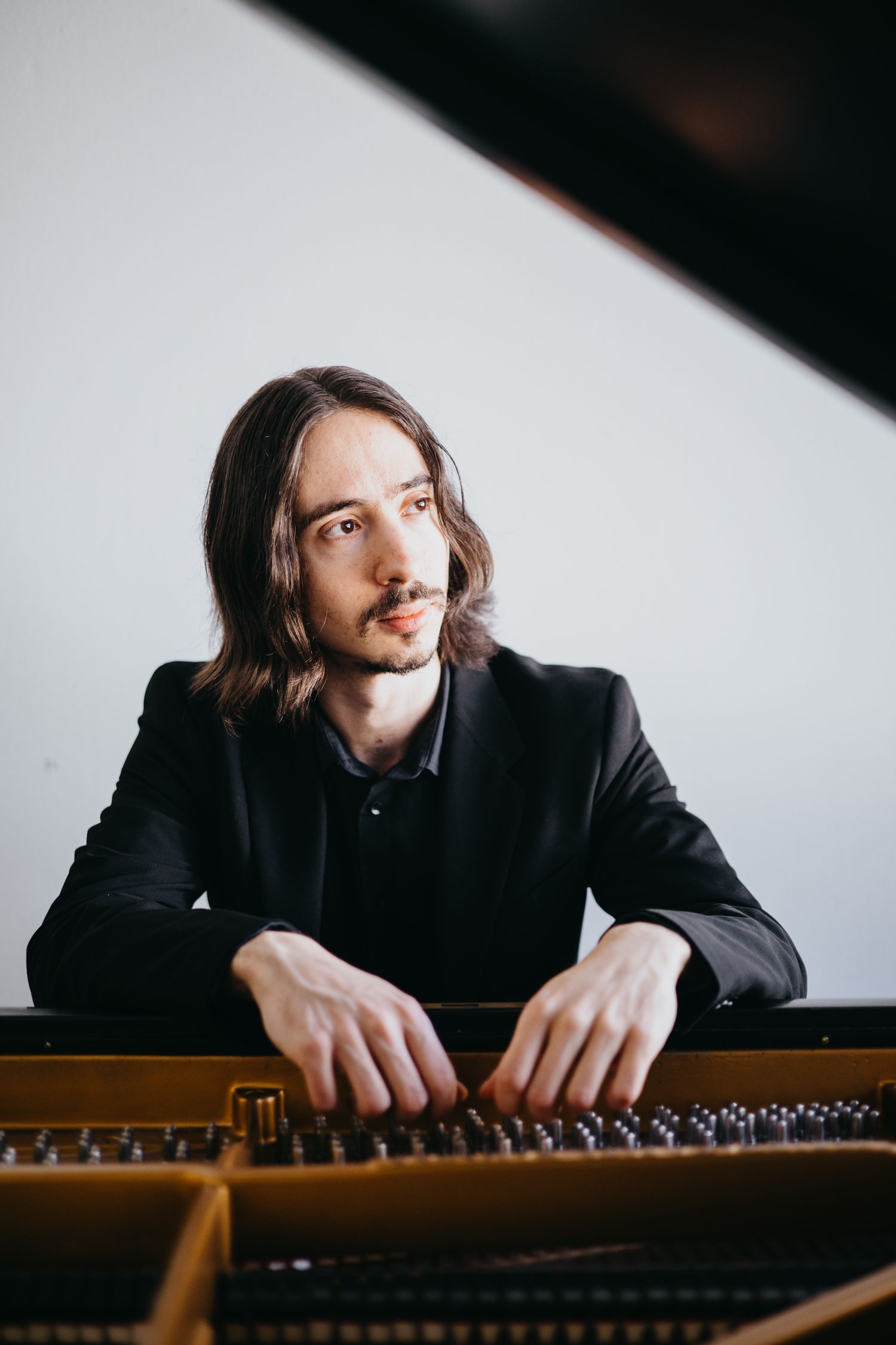 Long-haired male pianist in black suit seated at grand piano looking away.
