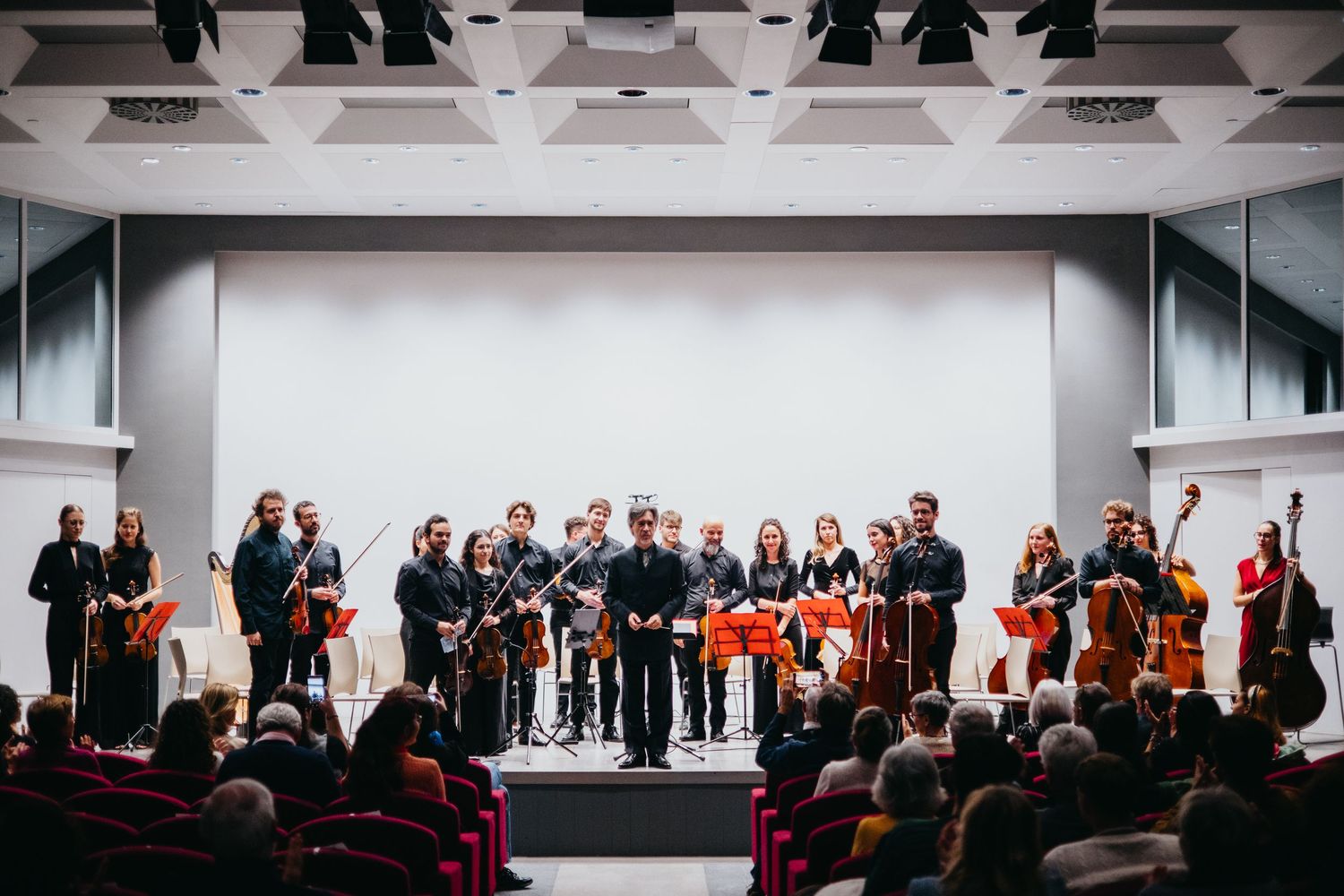 Chamber orchestra musicians bow on stage before seated audience in modern concert hall.
