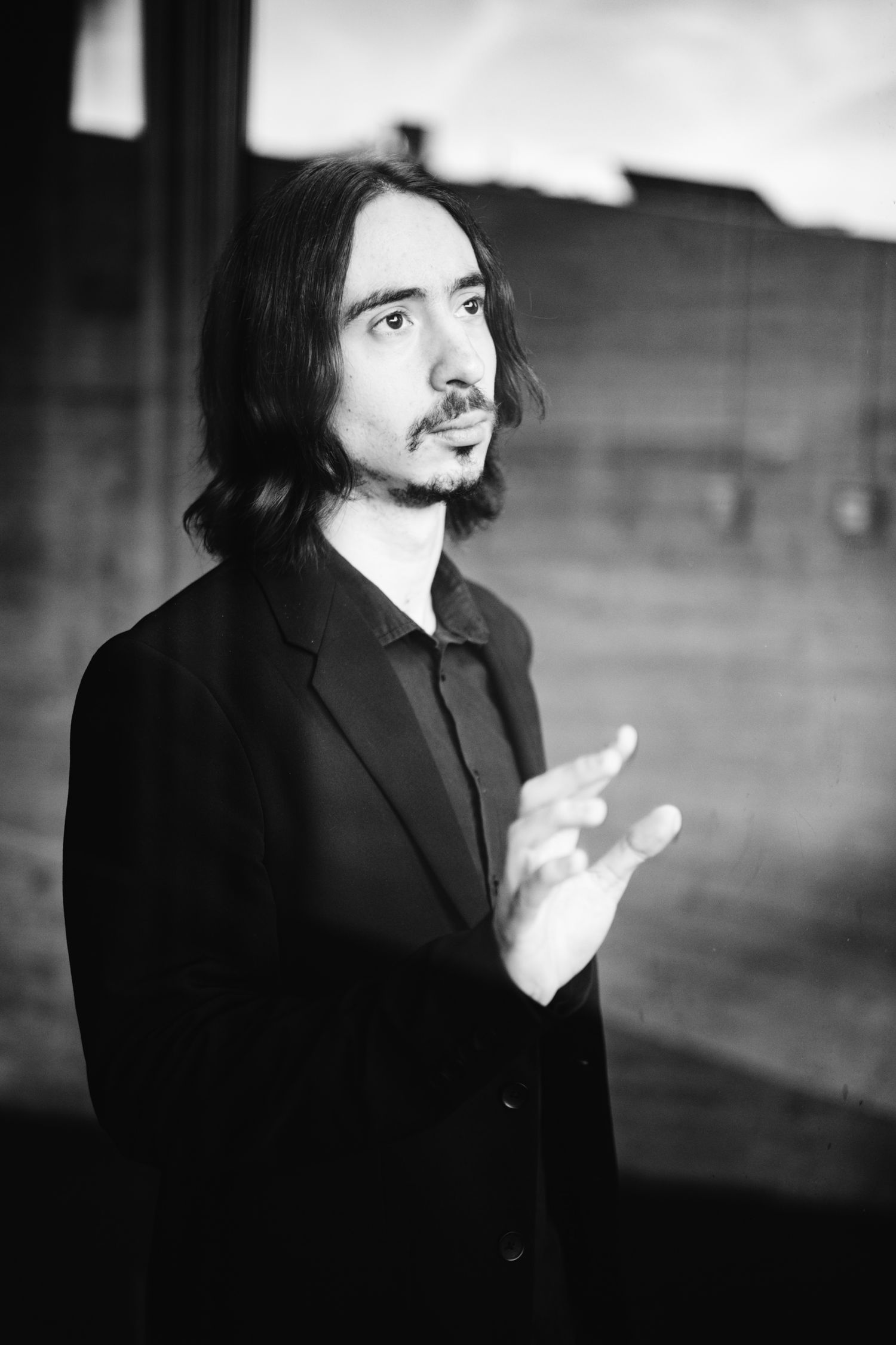 Black and white portrait of a long-haired musician in black suit gesturing with hand.