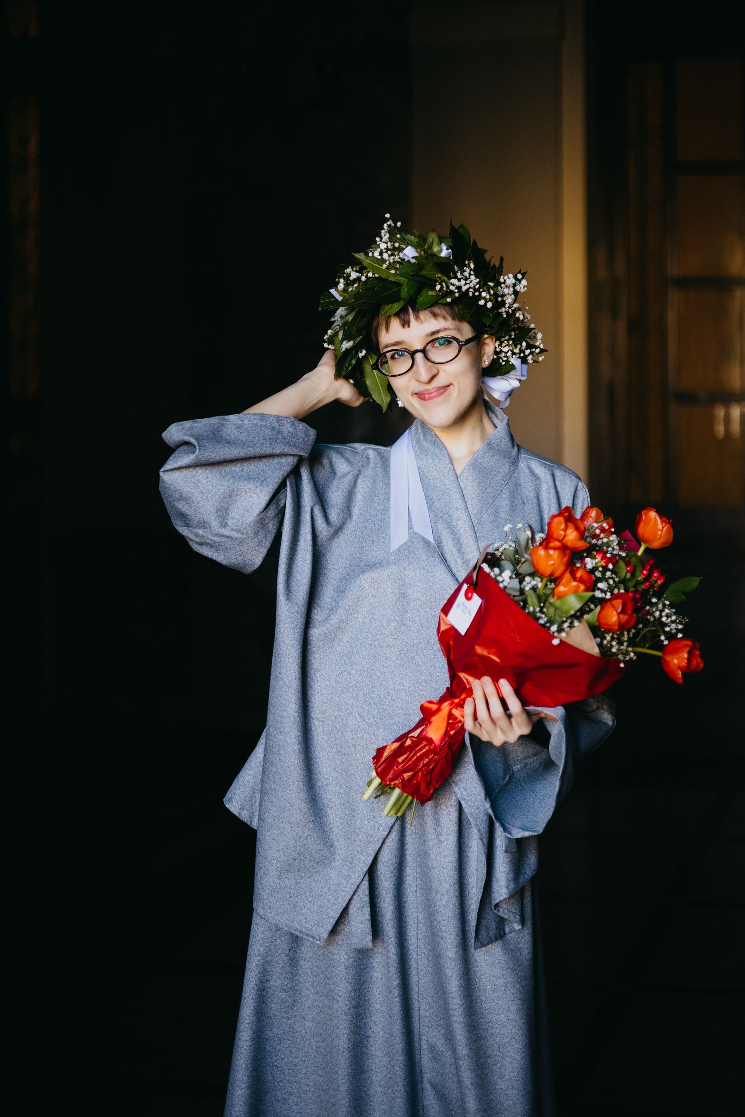 Smiling graduate in gray academic robe and floral crown holds vibrant red and orange bouquet.