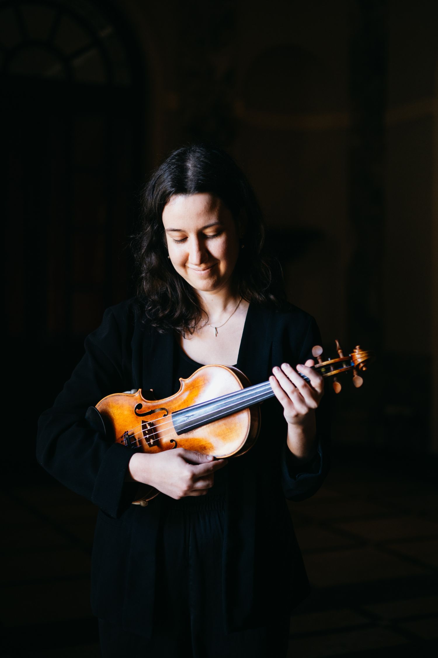 Young woman in black dress smiling while holding a violin in a dimly lit room.