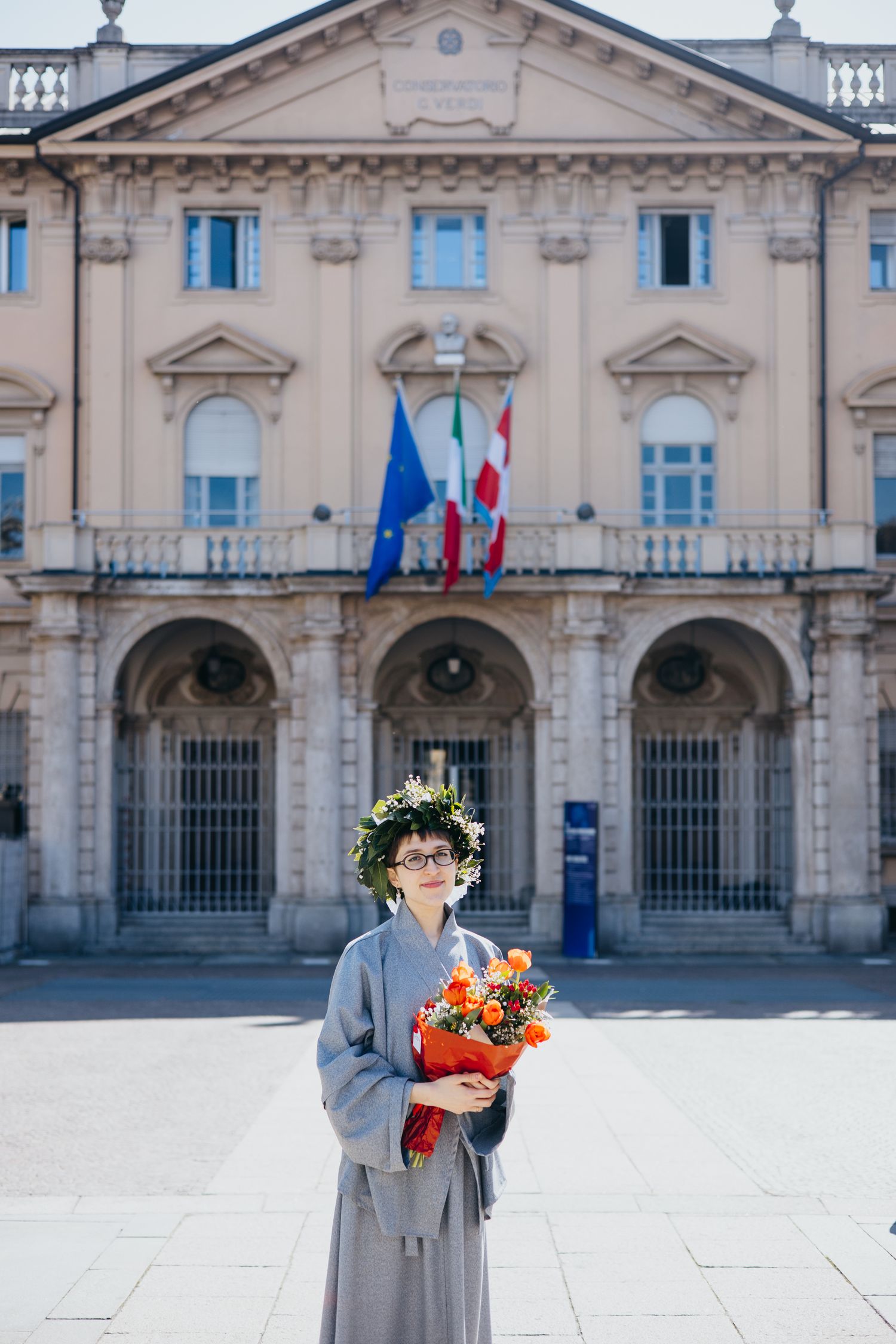 Graduate in gray robe and floral crown holds orange bouquet outside Italian municipal building.